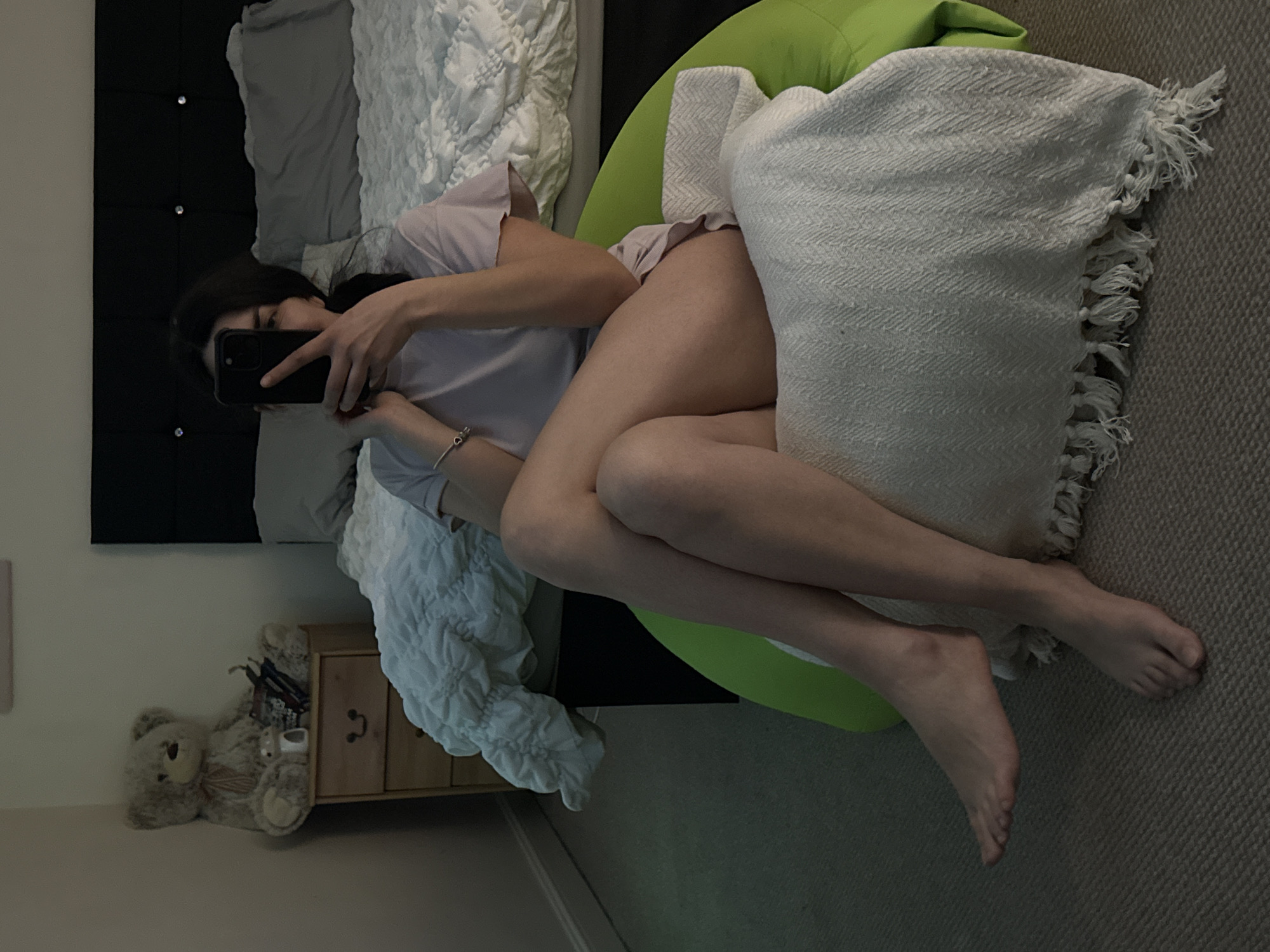 A young woman is sitting in a lime-green beanbag chair in a bedroom. She is holding a black phone, presumably taking a selfie. The woman is wearing a light-colored short-sleeved shirt and shorts or underwear. Her legs are crossed at the knees. A white blanket with tassels is draped over the beanbag chair and partially covers her legs.
Behind the woman is a black headboard with studded embellishments. A bed with white and gray bedding is visible behind the headboard. A small wooden nightstand is to the left of the bed. On the floor next to the nightstand is a teddy bear.
The room is dimly lit, and the floor is covered with a gray carpet.
The location is not discernible from the image and cannot be determined as Great Yarmouth, United Kingdom.