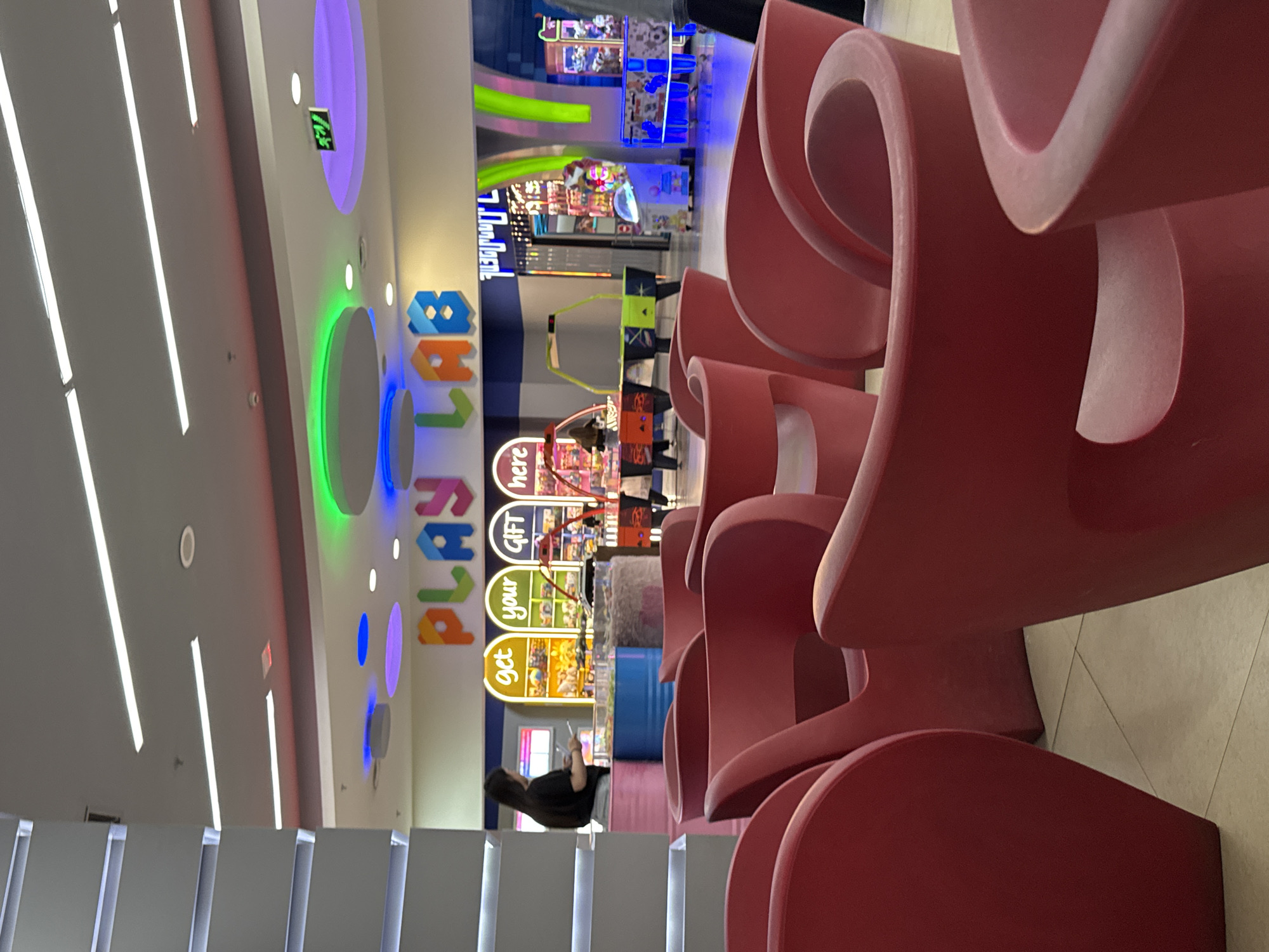 This image captures an indoor entertainment center or arcade, likely located in Yerevan, Armenia. The scene is brightly lit by a combination of white fluorescent tube lights on the ceiling and colorful LED lighting, including circular green and purple light rings.
In the foreground, a series of distinctive, sculptural red chairs or seating units with curved, wave-like designs are arranged. These modern furnishings lead the eye towards the main area of the entertainment center.
To the left (when viewed in corrected upright orientation), a person with dark hair is seated on a low white wall or bench, looking down at a mobile phone. This person is wearing a black top. Further back and to the right, numerous arcade games and attractions are visible, featuring bright screens and flashing lights typical of such venues.
A prominent sign on the wall reads "PLAY LAB" in large, colorful, block letters. Below and to the right of this, another vertical sign or booth is sectioned into three colorful, oval-shaped panels, each containing a word: "get," "your," and "GIFT," followed by "here" at the bottom. This indicates a prize or redemption counter. An illuminated green exit sign with a running man symbol is also visible on the ceiling. The overall impression is one of a vibrant, active, and modern leisure space.