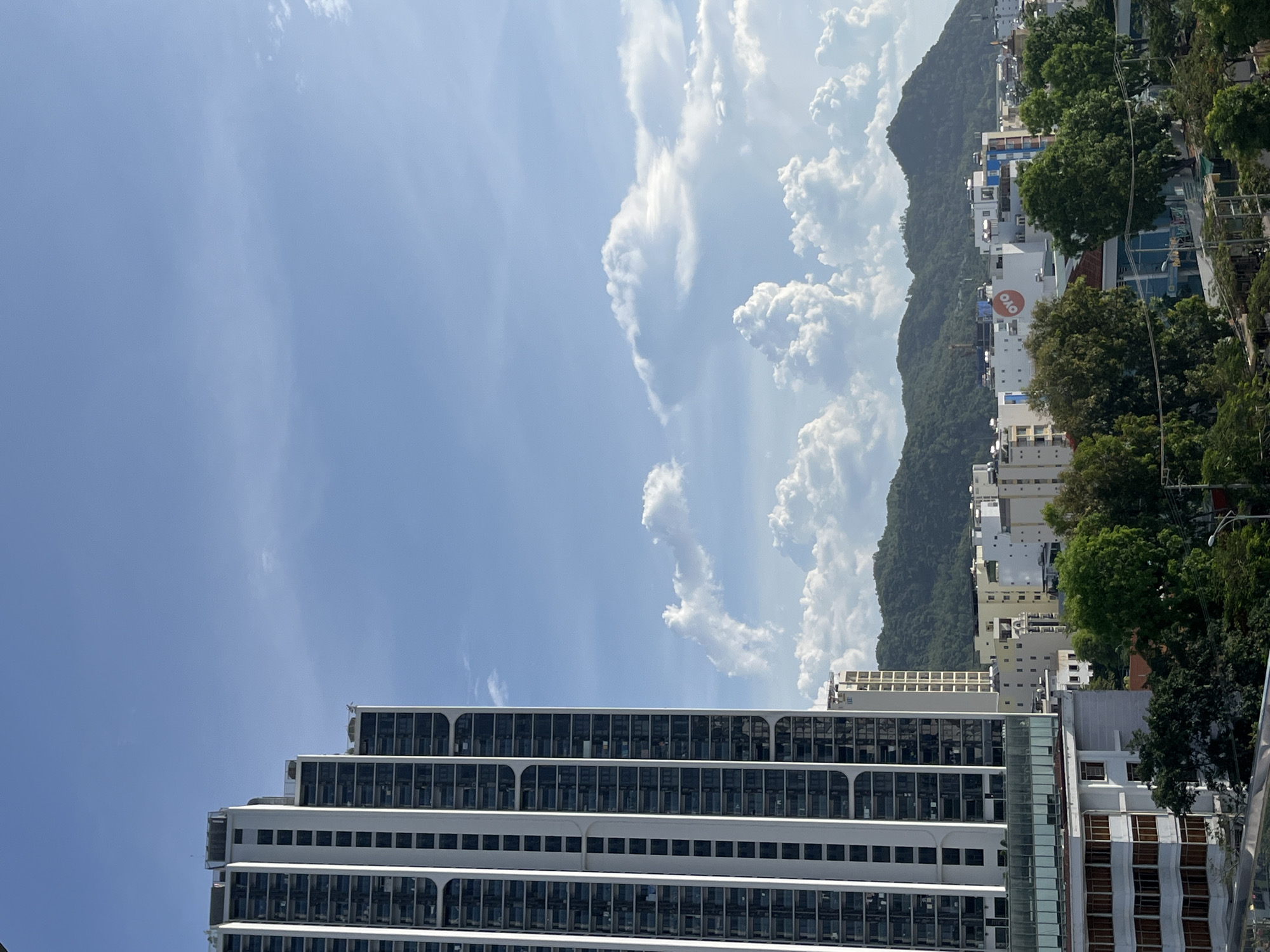 The image depicts a cityscape with a prominent modern high-rise building on the left, characterized by its white exterior and dark-tinted windows arranged in a grid pattern. To the right of this skyscraper, a range of smaller buildings and residential structures are visible, interspersed with lush green trees. In the background, verdant, rolling hills form a natural backdrop to the urban landscape. The sky above is a clear blue, dotted with fluffy white clouds, suggesting fair weather. The OYO logo is visible on one of the buildings in the mid-ground.