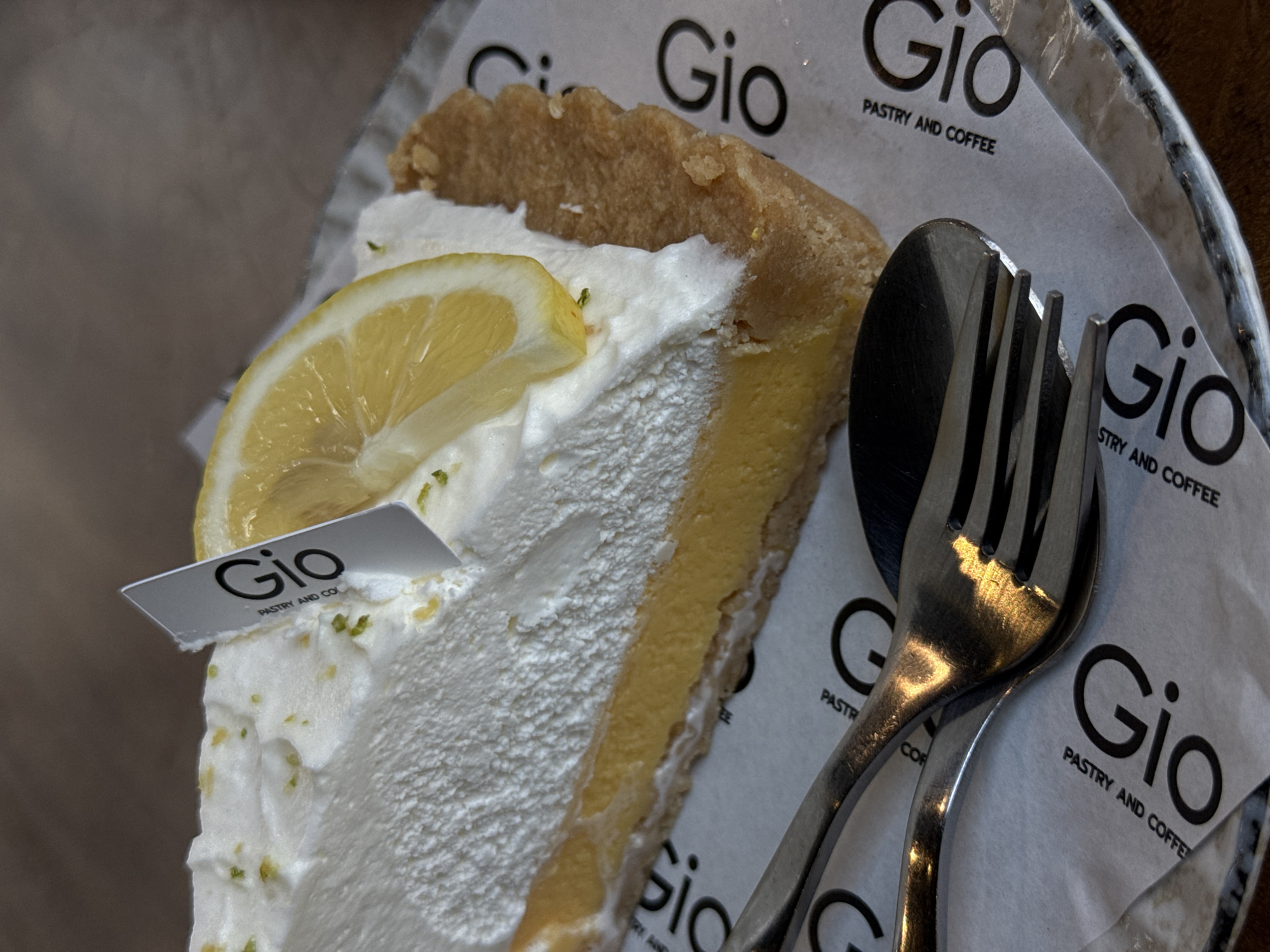 A close-up, eye-level, slightly angled shot shows a slice of lemon meringue pie on a white paper placemat. The pie has a golden-brown crust, a thick yellow filling, and a fluffy white meringue topping garnished with finely grated zest and a thin slice of lemon. A small, silver, rectangular tag with the black logo "Gio Pastry and Coffee" is inserted into the meringue. Two pieces of silverware, a fork and a spoon, are placed on top of the placemat, reflecting the overhead lighting. The background is a dark, textured surface, likely a wooden table. The placemat also features repeated "Gio Pastry and Coffee" logos. The image appears to have been taken in a cafe or restaurant setting in Baan Bo Phut, Thailand.