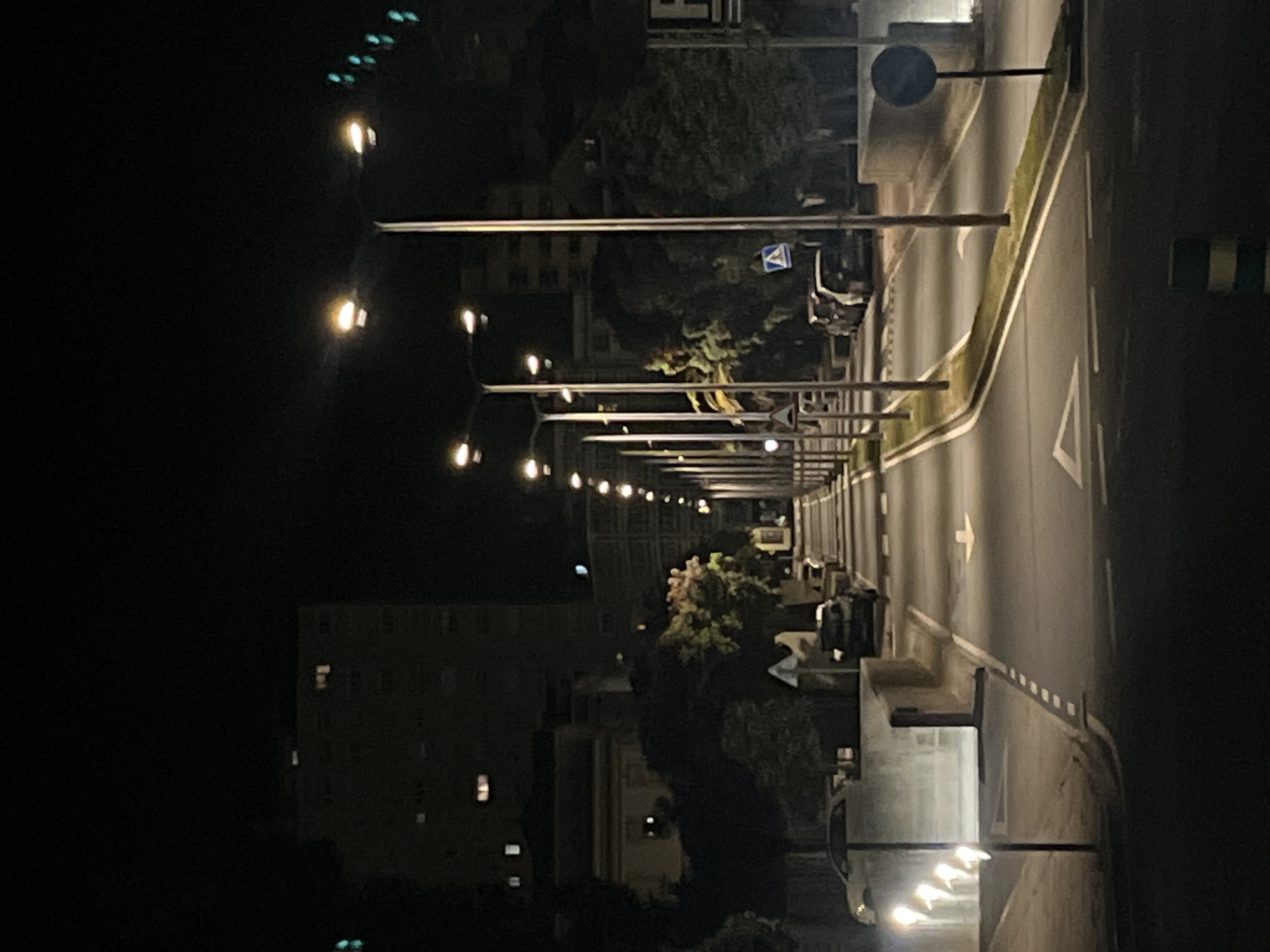The image shows a night scene along a street in A Coruña, Spain. The perspective is slightly elevated, looking down the street. The street is illuminated by a series of streetlights lining both sides. A multi-story building is visible on the left side of the frame with a few lit windows. On the right side of the street, there's a sidewalk and a road with lane markings, including arrows indicating direction. Trees and bushes are also visible along the street. The scene is mostly dark due to the nighttime setting, with the streetlights and building windows providing the primary sources of illumination. A few cars can be seen parked along the street. Road signs are also visible near the side of the road.