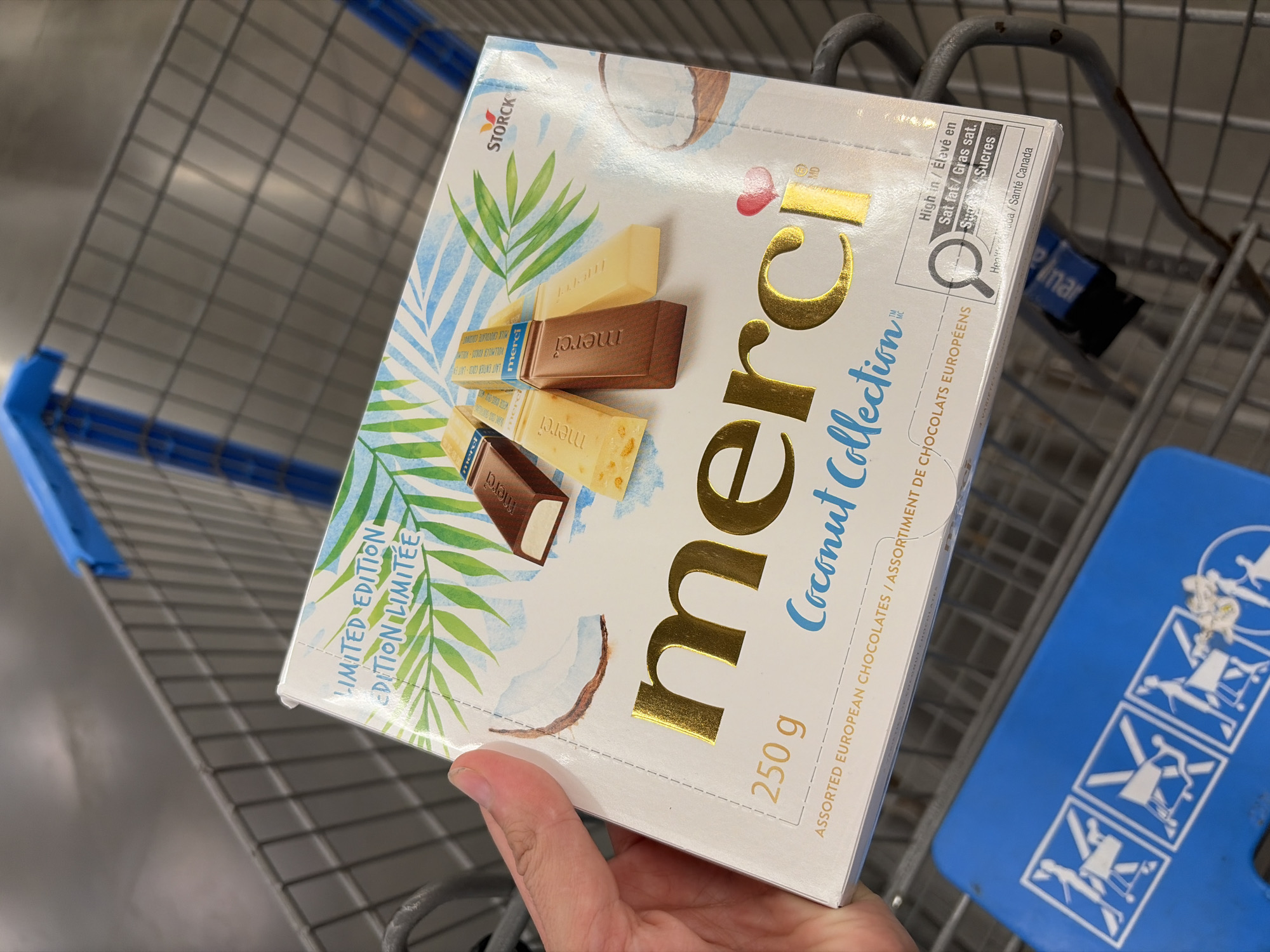 In an indoor setting, likely a retail environment such as a grocery store, a hand holds a box of Merci Coconut Collection assorted European chocolates. The box is white with a tropical design featuring palm leaves and images of coconut and chocolate bars. Text on the box indicates "LIMITED EDITION," "250 g," and "Coconut Collection." Below this, it reads "ASSORTED EUROPEAN CHOCOLATES / ASSORTIMENT DE CHOCOLATS EUROPÉENS." To the right, a nutrition label highlights "High in / Élevé en Sat fat / Gras sat." and "Sugar / Sucres." In the background, the wire frame of a shopping cart is visible. A blue handle of a shopping cart is also visible in the foreground.