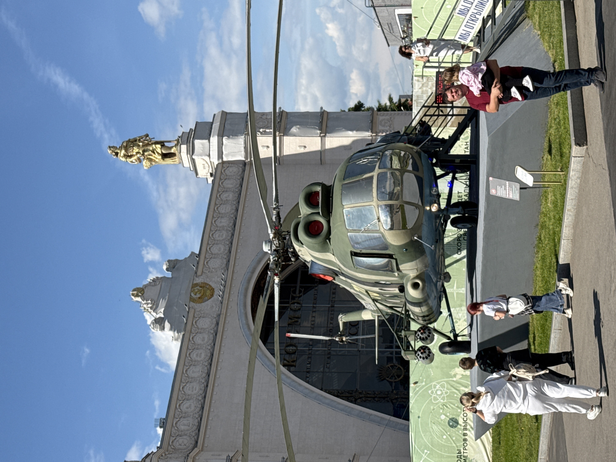 An outdoor scene in Moscow, Russia, features a large military helicopter on display in front of a grand, ornate architectural structure under a bright blue sky with scattered clouds. The image captures a sunny daytime setting.
In the foreground, a large, olive-green helicopter, likely a Mil Mi-8 or Mi-17 series, is exhibited. Its main rotor blades are visible, extending upwards. The helicopter's cockpit windows are reflective, and visible elements include external missile pods and landing gear. It appears to be part of an outdoor exhibition, with informational signs placed near it.
Behind the helicopter stands a monumental building, characterized by its classical architecture, intricate carvings, and columns. A golden statue of a figure holding a sheaf of wheat, possibly a representation of a worker or collective farmer, tops a prominent part of the building. The building's facade also features circular decorative elements and what appear to be symbols of the hammer and sickle, consistent with Soviet-era architecture often found at locations like VDNKh.
Several people are present, observing the helicopter and the surroundings. On the right, a man in a red shirt holds a young child in a pink top, both looking towards the helicopter. Further back, another woman in a white dress stands near a banner. Closer to the foreground, a group of people, including individuals in a white tracksuit and others in casual wear, are also seen.
Visible text on various banners and signs includes:
- "КОМОС"
- "МЫ О" (likely part of "МЫ ОТКРЫЛИ" - "We opened")
- "МЕТРОВ В ВЫСОТУ" ("meters in height")
- "СКОРО" ("soon" or "speed")
- "ДЕТ КАЛЬНЫ" (potentially part of "уникальный" - "unique")
- "TAH" (possibly part of a longer word like "выставка" - "exhibition")
The scene depicts a public event or museum visit, where visitors are engaging with historical or technological exhibits against a backdrop of significant architecture.