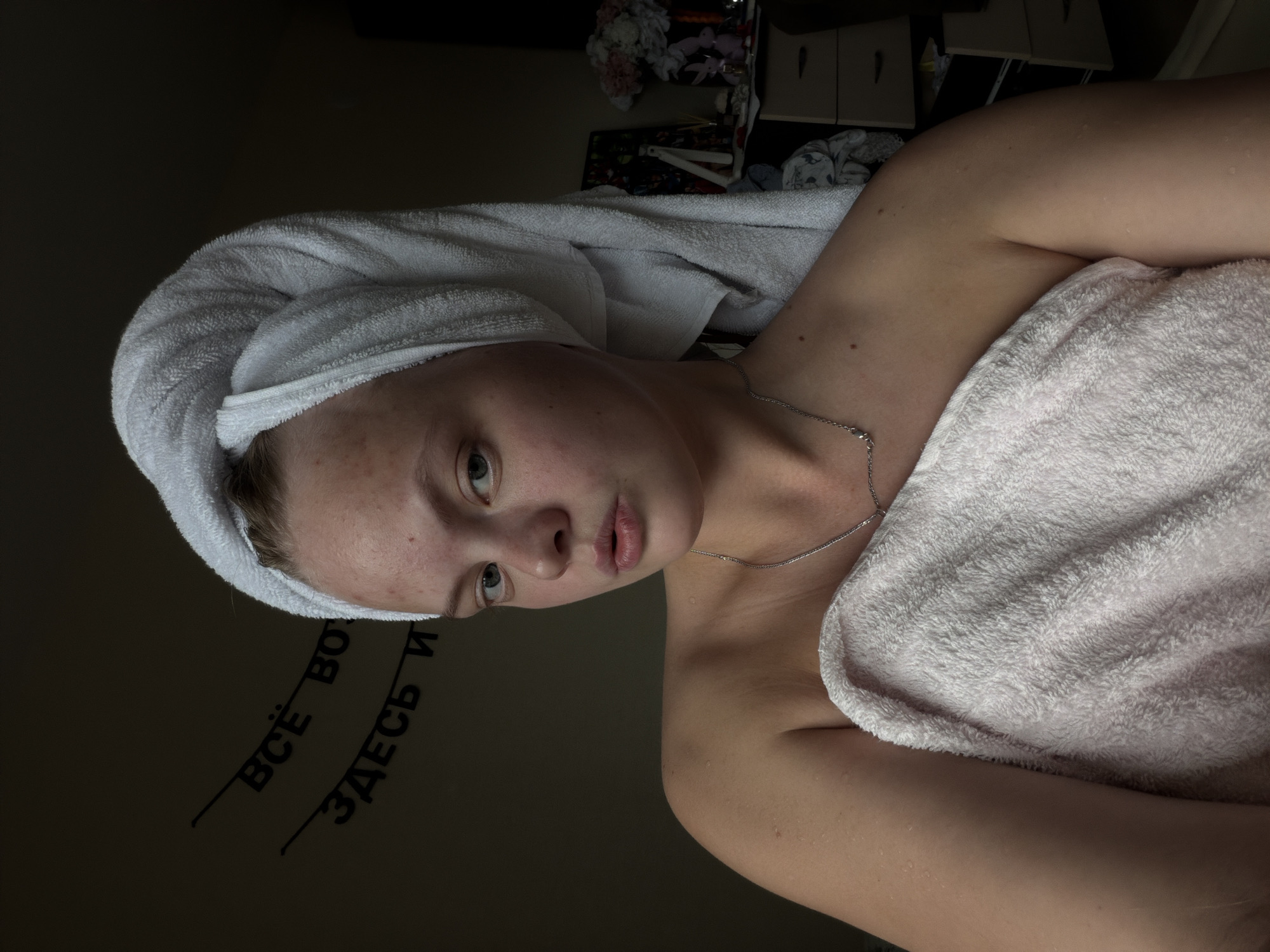 The image features a young individual, likely female, captured in a dimly lit indoor setting, possibly a bedroom or bathroom. The person's body is wrapped in a light pink or white towel, and their head is wrapped in a white bath towel, suggesting they have recently showered or bathed. Their skin appears fair with some freckles and small water droplets visible on the shoulder and chest, enhancing the post-bath impression. They wear a simple silver chain necklace.
The individual is looking directly towards the camera with a neutral expression, lips slightly parted. The photograph is taken from a slightly high angle, and the image itself is rotated counter-clockwise.
The background is dark and largely out of focus, but some details are discernible. On the upper right, there are shelves or drawers with various items, including what appears to be decorative flowers and a small object resembling a balisong (butterfly knife) placed on a shelf. To the lower left, on a wall or surface, there is visible text in Cyrillic, rotated along with the image. The legible parts read "ВСЁ ВО" (VSE VO) and "ЗДЕСЬ И" (ZDES' I), which translates to "EVERYTHING HERE AND...". The overall lighting is soft and subdued, creating an intimate atmosphere. The location context of Izhevsk, Russia, is consistent with the visible Cyrillic text.