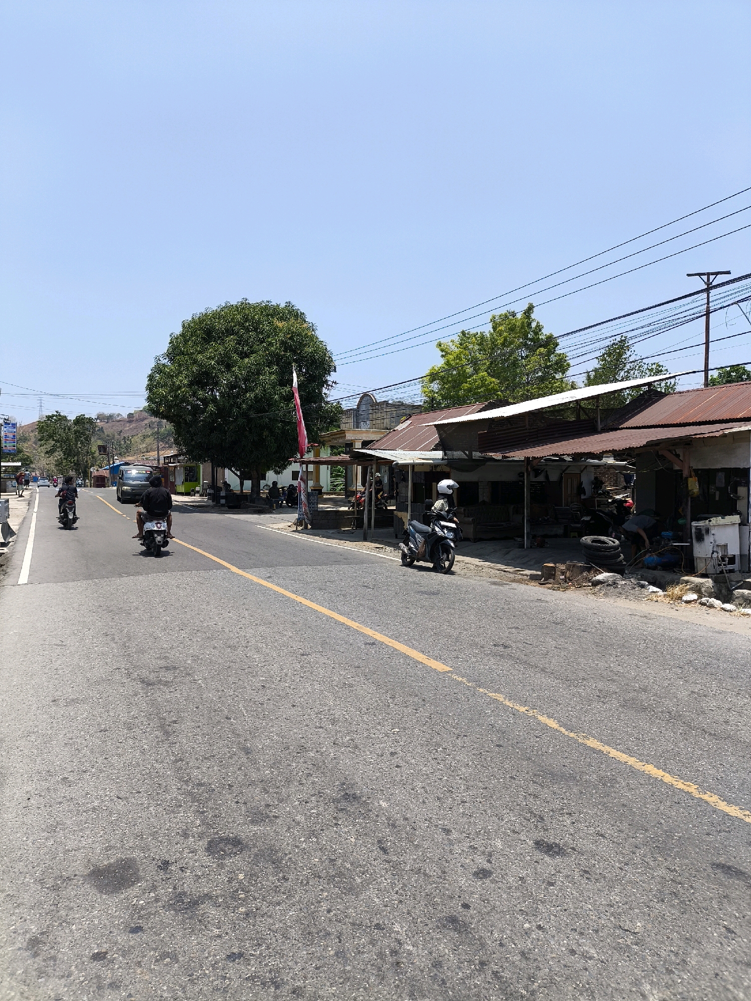 An eye-level, medium shot captures a two-lane road in a village-like area of Unknown, Indonesia on a clear, sunny day. The asphalt road, marked with a single yellow line down its center, is in the foreground and middle ground. To the right of the road, a row of small, rustic buildings with corrugated metal roofs are visible, appearing to be shops or workshops. One building has several tires stacked outside, and a person is working on machinery within the open-fronted structure. A scooter with a rider wearing a helmet is parked near the front of this building. Further down the road on the right, more structures and trees are present.
On the left side of the road, a large, leafy green tree stands prominently. Beyond the tree, a slight incline with sparse vegetation and some distant buildings can be seen, suggesting the edge of the village. A few people on motorcycles are traveling along the road. One motorcycle is in the foreground on the left, with a rider wearing a black shirt. Another motorcycle is further ahead on the left, also with a rider in a dark shirt. A van or truck is parked in the distance on the left side of the road, and some utility poles and power lines stretch across the sky. The sky is a clear, bright blue.