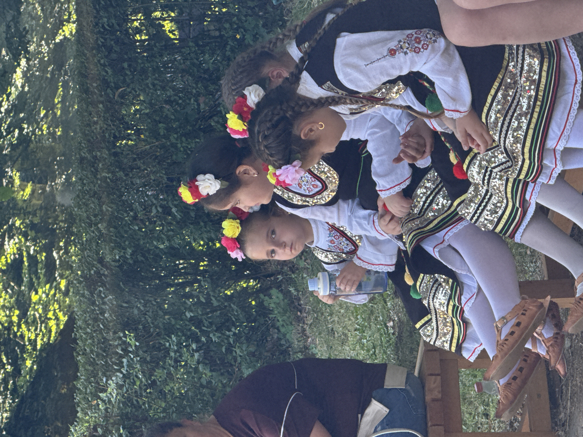 In Ribaritsa, Bulgaria, several young girls in traditional Bulgarian folk costumes sit on a wooden bench, facing left. They are adorned with floral headbands featuring red, yellow, and white flowers, and wear embroidered black vests over white blouses with red and white trim. Their skirts are black with gold sequined trim and red and green accents, worn over white tights. The girl in the foreground on the right has braided dark hair and wears a gold hoop earring. The girl in the middle is holding a blue and white water bottle. Their feet are clad in brown, laced moccasin-style shoes. Behind them, a dense green hedge forms the backdrop, with dappled sunlight filtering through trees. To the left, an adult's arm and shoulder are visible, wearing a dark maroon shirt.
