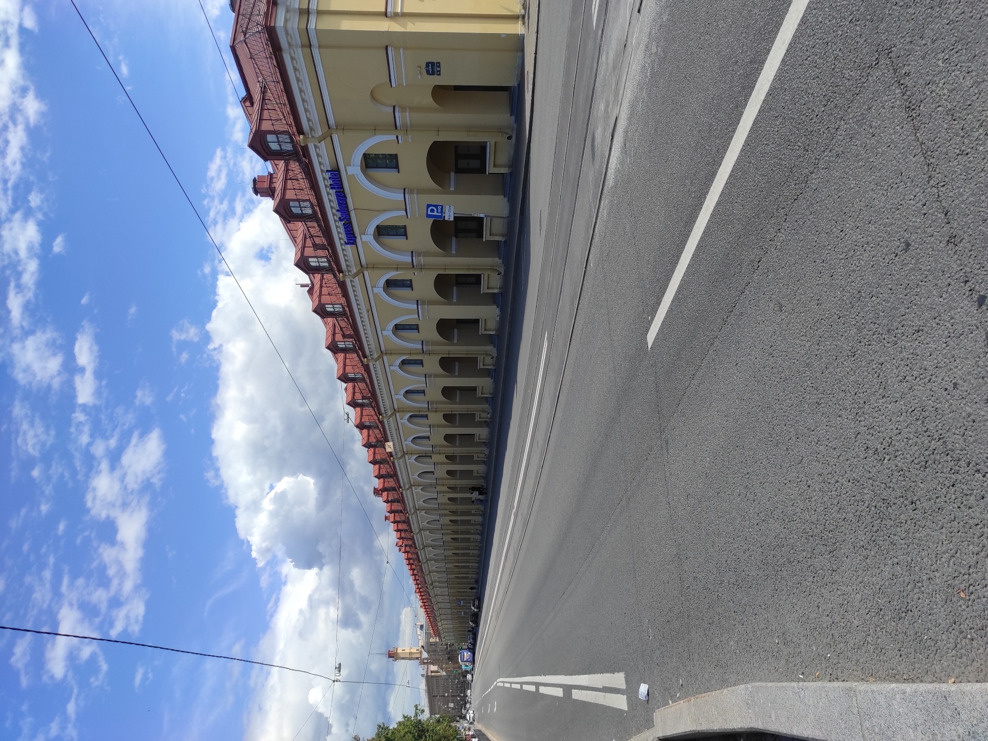 The image shows a view of the Nevsky Prospekt in Saint Petersburg, Russia, on a sunny day with scattered clouds. A long, yellow building with a red-tiled roof and arched colonnades runs along the right side of the street. A blue sign on the building reads "Express Sadovaya Hotel". A blue bus is visible in the distance on the left side of the street, along with several parked cars and some pedestrians walking in front of the hotel. The street is paved with asphalt and has tram tracks running along its length. A white line marks the lane on the right side of the road. A grey curb runs along the left side of the street, with some litter on the pavement. In the far distance on the left, a tall, light-colored tower with a red spire is visible.