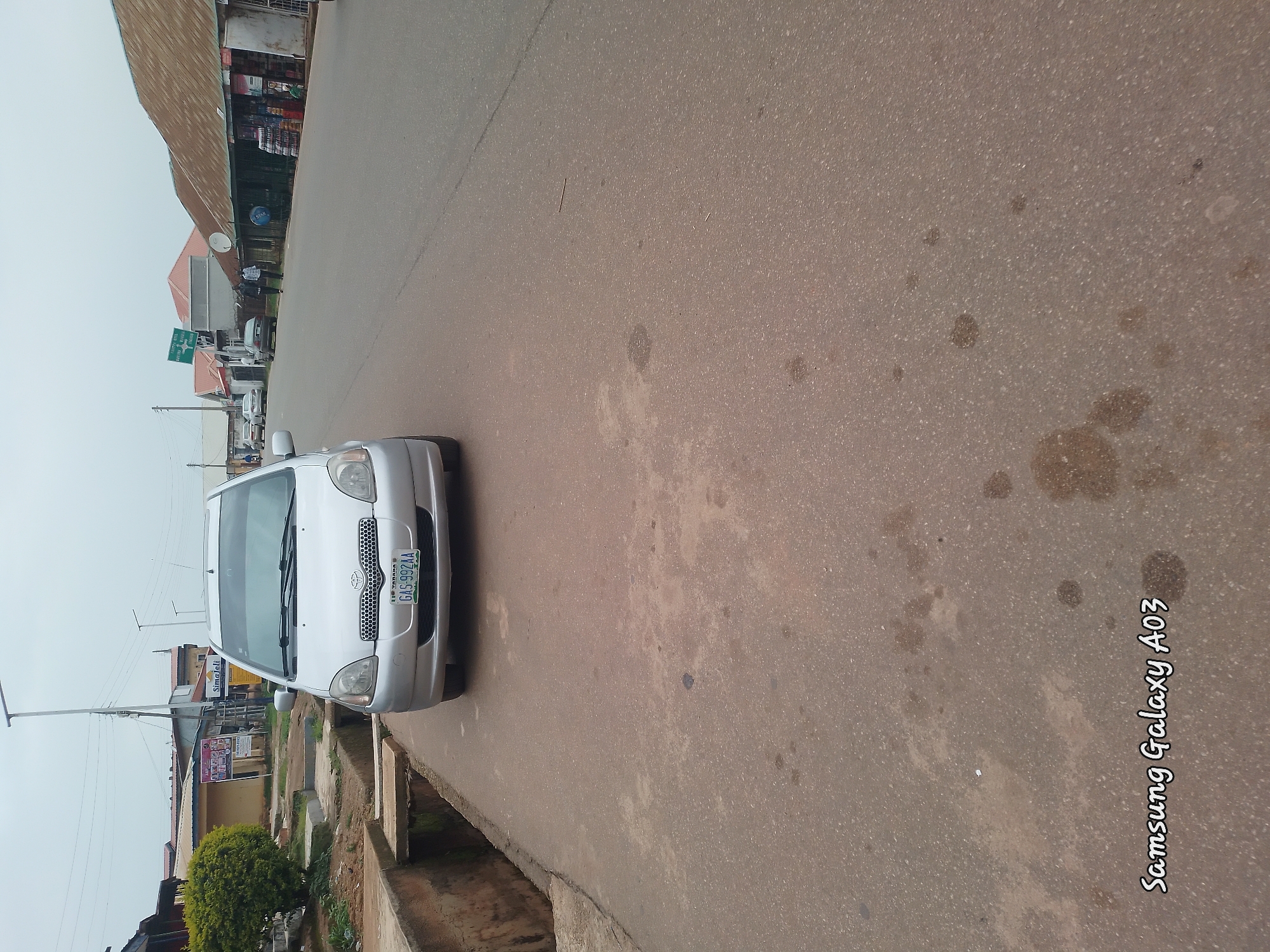 The image shows a silver Toyota Yaris parked on the side of a road in Jos, Nigeria. The car is facing forward, and its license plate reads "GAS-992AA". The road appears to be in a commercial area, with several shops and buildings lining the street. There are also a few people walking in the background. The sky is overcast, and the overall lighting is dim. The image also contains a watermark that says "Samsung Galaxy A03" in the bottom left corner.