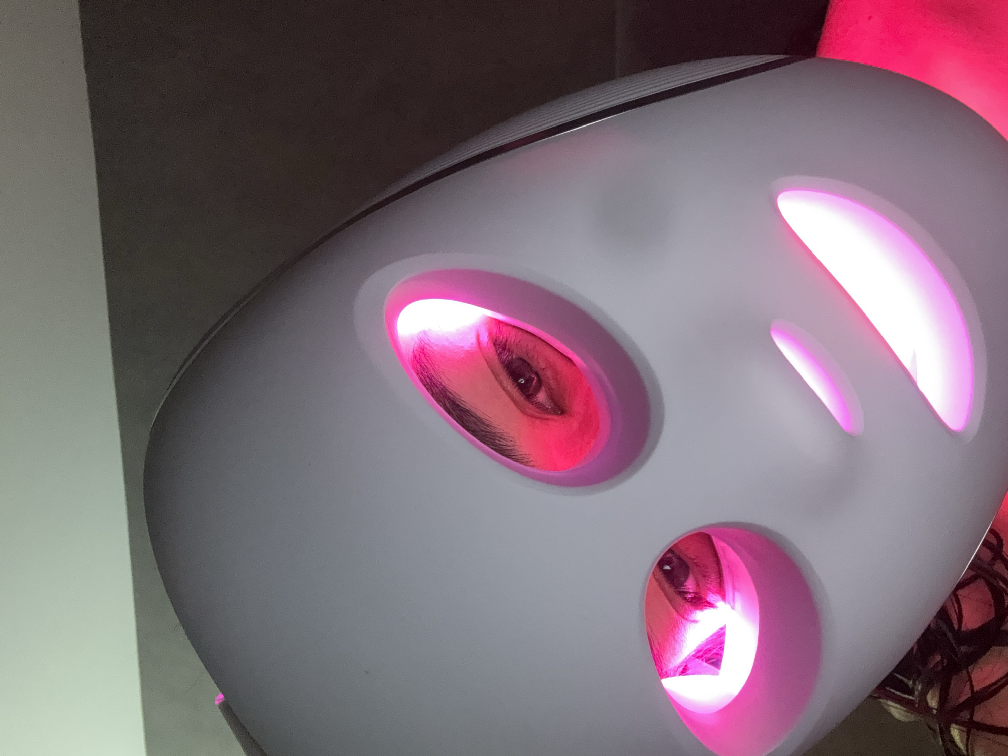 The image captures a close-up perspective of a person undergoing a cosmetic or therapeutic treatment using an LED light therapy mask. The main subject is partially visible through the mask, specifically their eyes and eyebrows.
The prominent object is a sleek, white or light gray mask-like device that covers the face. This device features several oval-shaped openings, two of which align with the person's eyes, allowing them to look out. The interior of the mask and the areas around the eyes are brightly illuminated with a vibrant pink or magenta light, suggesting a specific wavelength of light therapy, often associated with red light.
Through the eye openings, the person's left eye and part of their right eye are clearly visible. They have dark eyebrows and eyelashes, and their eyes are open, looking directly forward. The skin around the eyes is bathed in the pink light.
The background is indistinct, appearing as a dark gray or black surface on the upper right and a lighter gray or white surface on the left, possibly walls in an indoor setting. The image is taken at an oblique angle, emphasizing the device and the illuminated eyes. No visible text is present in the image. The implied activity is a beauty or wellness treatment, possibly for skin rejuvenation or therapy. The location context provided is San Bernardino Tlaxcalancingo, Mexico, though no visual cues confirm this.