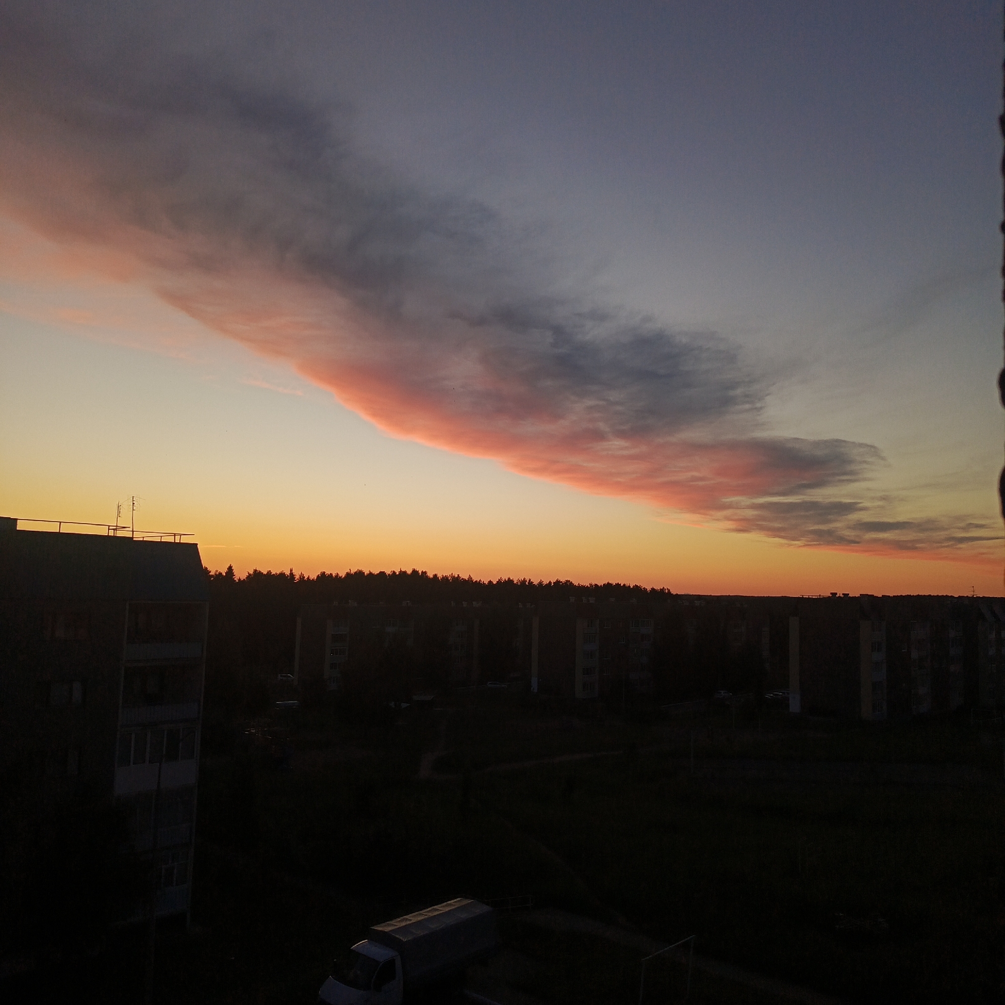 The media file captures a serene sunset or sunrise over a residential area in Markovsky, Russia. The sky is the dominant subject, featuring a magnificent, elongated cloud formation stretching horizontally across the upper half of the frame. This cloud displays vibrant hues of pink and orange at its lower edge, transitioning into darker blues and grays towards its upper parts, contrasting with the clear, bright band of orange and yellow light just above the horizon. The sun itself is not visible, having either just set or not yet risen above the horizon line.
Below the colorful sky, the landscape is rendered in deep silhouette. A dense line of dark trees forms a natural horizon, behind which several multi-story apartment buildings are visible. These buildings, typical of Soviet-era architecture, appear as dark masses against the fading light, with some window details faintly discernable upon closer inspection. In the bottom-left, a portion of another apartment building is more clearly outlined, showing balconies and windows.
In the foreground, a white truck, possibly a delivery vehicle, is parked and partially illuminated by the faint ambient light, appearing in the bottom-middle. On the rooftop of the building in the middle-left, a small antenna structure is visible. No people are present in the image, and there is no visible text. The overall atmosphere conveyed is peaceful and dramatic, highlighting the natural beauty of the sky at twilight over an urban landscape.