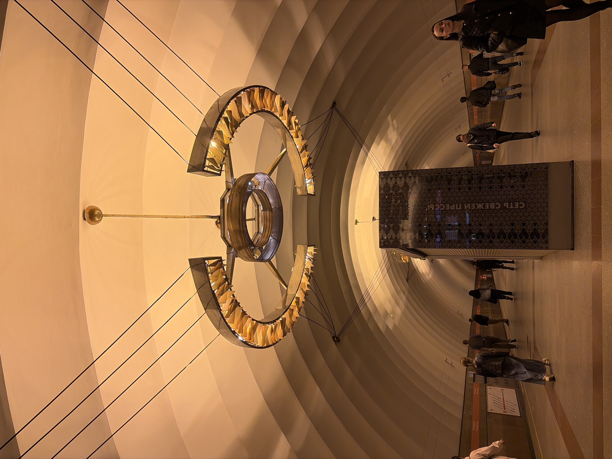 The image depicts a station of the Saint Petersburg Metro in Russia. In the foreground, a large, ornate chandelier-like structure hangs from the ceiling. It consists of a central ring with a vertical post extending from the top, flanked by curved metallic elements adorned with smaller, blade-like forms. Supporting cables connect the chandelier to the arched ceiling.
The station's architecture features a curved tunnel design with concentric rings of light and shadow. On the right side of the image, a platform is visible with several individuals standing and walking. A dark rectangular structure, possibly an information kiosk or advertising display, is positioned near the edge of the platform. A text inscription, "СЕТЬ СВЕЖЕЙ ПРЕССЫ", is visible on it. People are either standing waiting, or walking by.