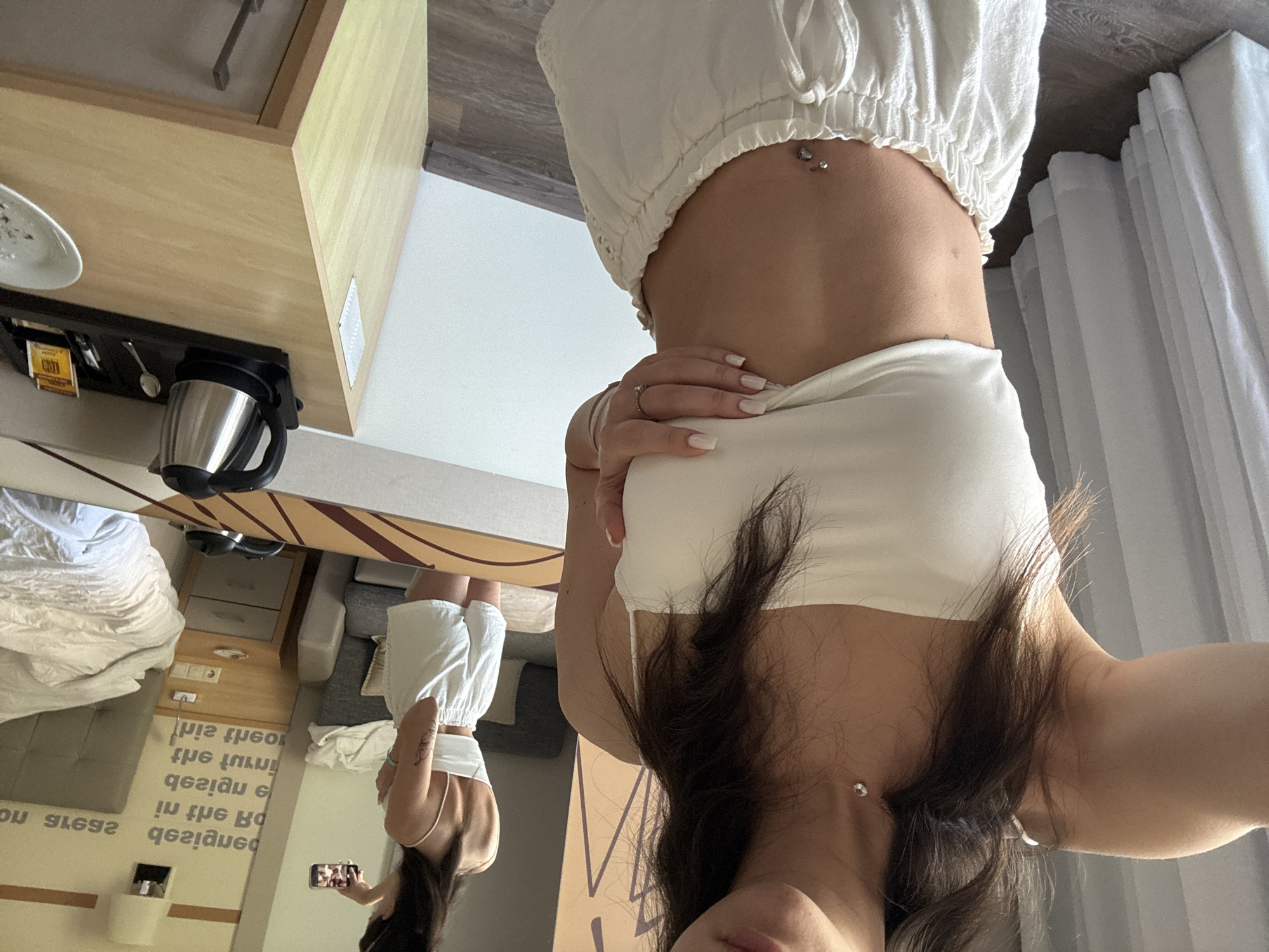 A young woman with dark hair is taking a selfie in a hotel room. She is wearing a white crop top and white shorts. She has a belly button piercing. In the background, a mirror reflects her and the room. The room contains a bed, a nightstand with a kettle, and a chair. The walls are decorated with a large, abstract pattern.