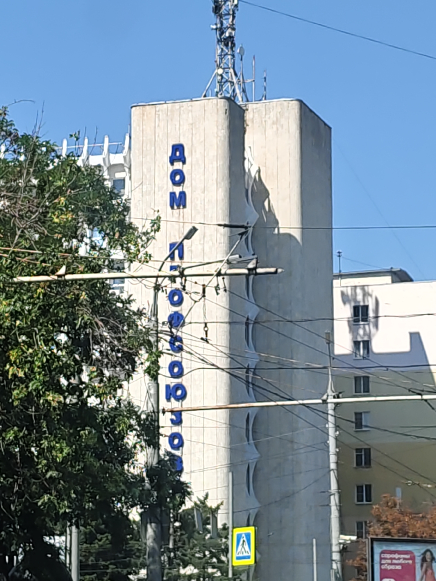 The image shows a tall, concrete building with the word "ДОМ" (DOM) in large blue letters vertically displayed on its facade. Below "ДОМ", additional blue letters are visible, reading "КПРФ" (KPRF), spelling out "Дом КПРФ", which translates to "House of the Communist Party of the Russian Federation". The building has a distinctive, wavy architectural feature running down its side. A telecommunications tower is mounted on top of the building. Electrical wires and poles are prominent in the foreground, and a pedestrian crossing sign is visible on the left. Another building, colored yellow, is seen to the right of the main structure, with windows and balconies. Trees with green foliage are present on the left side of the image. The scene appears to be a street view in a city on a clear, sunny day. This building is located at 48 Prospekt Budyonnovsky, Rostov-on-Don, Russia.