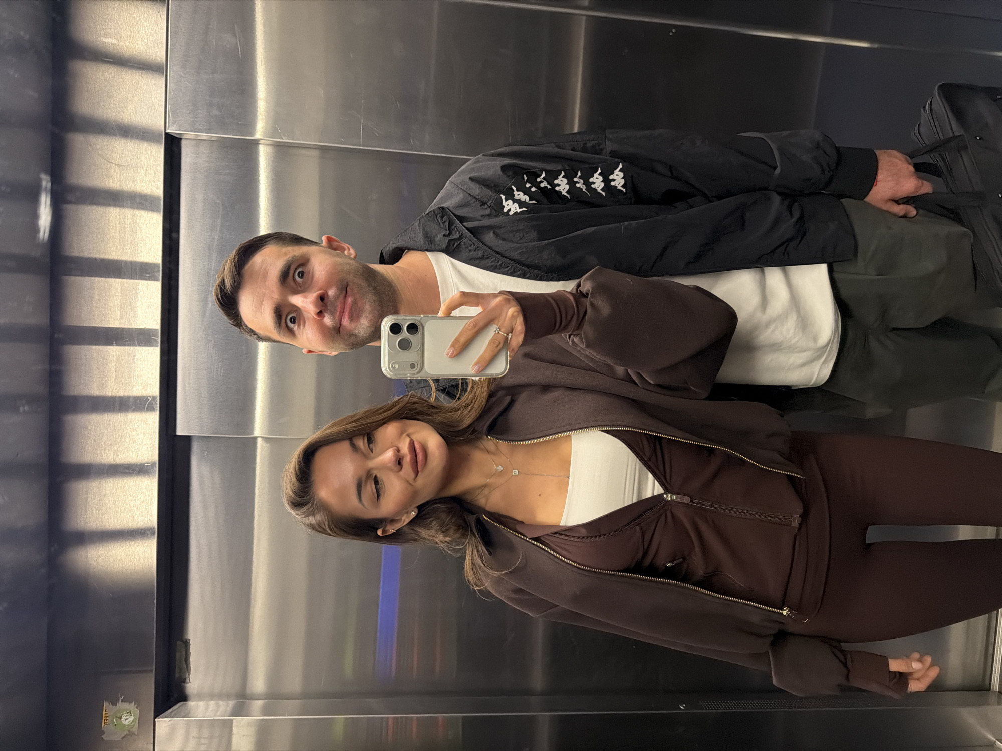 A mirror selfie captures a man and a woman inside an elevator in Krasnoyarsk, Russia. The image is oriented vertically, showing the subjects reflected in the polished metallic walls.
On the right side of the frame, a man with short dark hair and light stubble looks towards the camera with wide, expressive eyes and a slight smirk. He wears a black jacket adorned with several white Kappa logos along the sleeve, over a white t-shirt, and dark-colored pants.
On the left, a woman with long brown hair holds a light-colored iPhone in a clear case to take the photograph. She is wearing a dark brown zip-up hoodie over a white bandeau top, paired with matching dark brown leggings. Her expression is a soft smile with a playful wink. She accessorizes with a ring on her left ring finger and a necklace featuring two small square pendants.
Behind the man, a black bag is visible. The elevator's walls are made of reflective brushed metal, showing faint reflections of ambient light and architectural lines. A small, illegible sticker is visible on the lower left wall. The overall atmosphere is casual and cheerful.