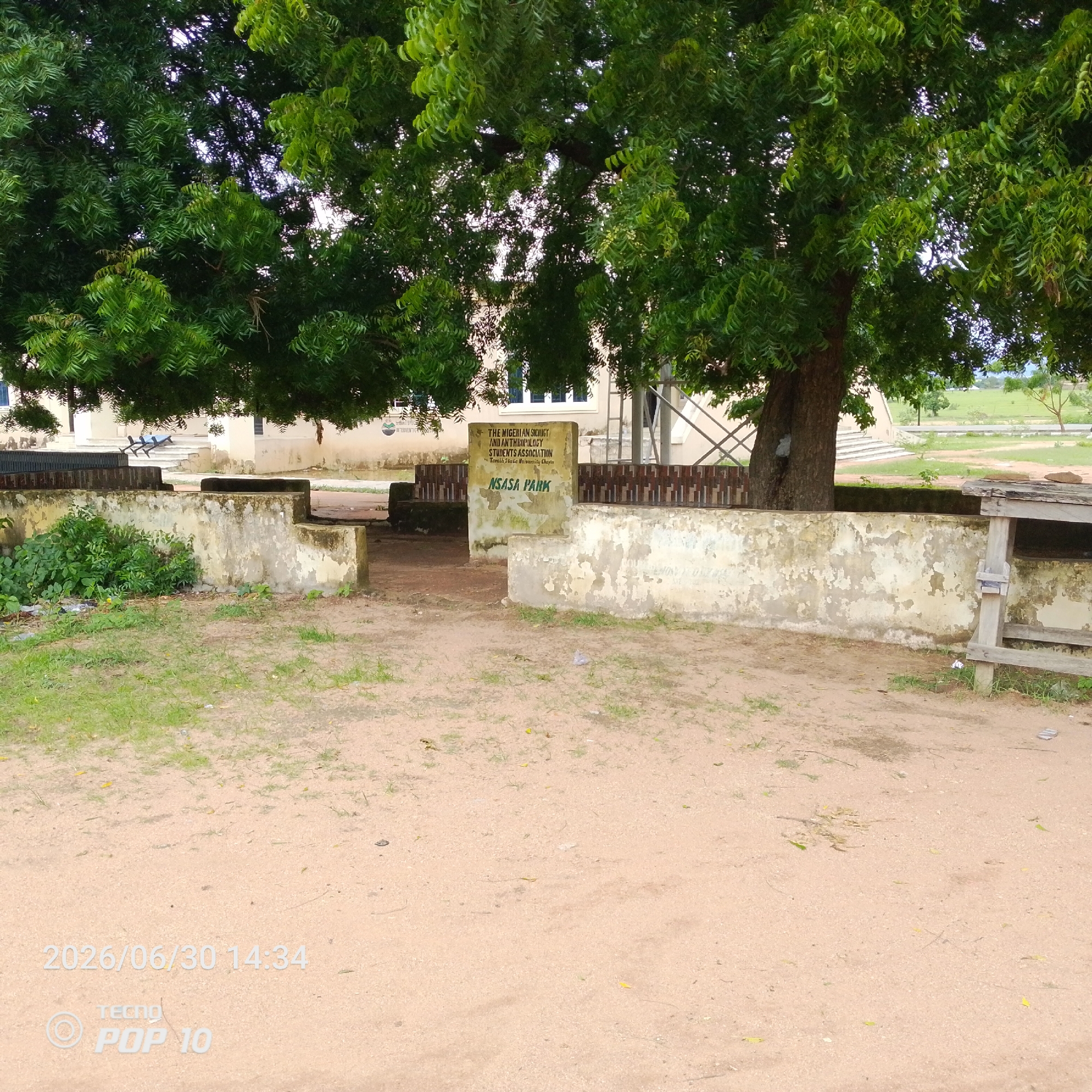 The image captures an outdoor scene in Jalingo, Nigeria. The primary subject is a weathered concrete structure, part of what appears to be an entrance or seating area, covered by the lush green foliage of a large tree. The tree's branches extend over the scene, creating dappled shade. Behind the concrete structure, a building with windows is visible, along with a metal fence.
The concrete structure features a sign with text. The visible text reads: "THE NIGERIAN SOCIOLOGY AND ANTHROPOLOGY STUDENTS ASSOCIATION Taraba State University Chapter" and below it, "NSASA PARK". This indicates the location is a park associated with a university student group.
The ground is a mixture of dirt and patches of dry grass. There are some scattered leaves and small debris on the ground, suggesting it's an outdoor, possibly natural or semi-maintained environment. The lighting suggests it is daytime, with the sun filtering through the tree canopy.
In the lower right corner of the image, a portion of a wooden structure, possibly a table or bench, is visible. The bottom of the image also contains visible text overlay: "2026/06/30 14:34 TECNO POP 10", which appears to be a timestamp and device watermark from a photograph taken with a Tecno Pop 10 phone.
Overall, the image depicts a peaceful, somewhat rustic park area at a Nigerian university, marked by a significant tree and a student association's designated space.