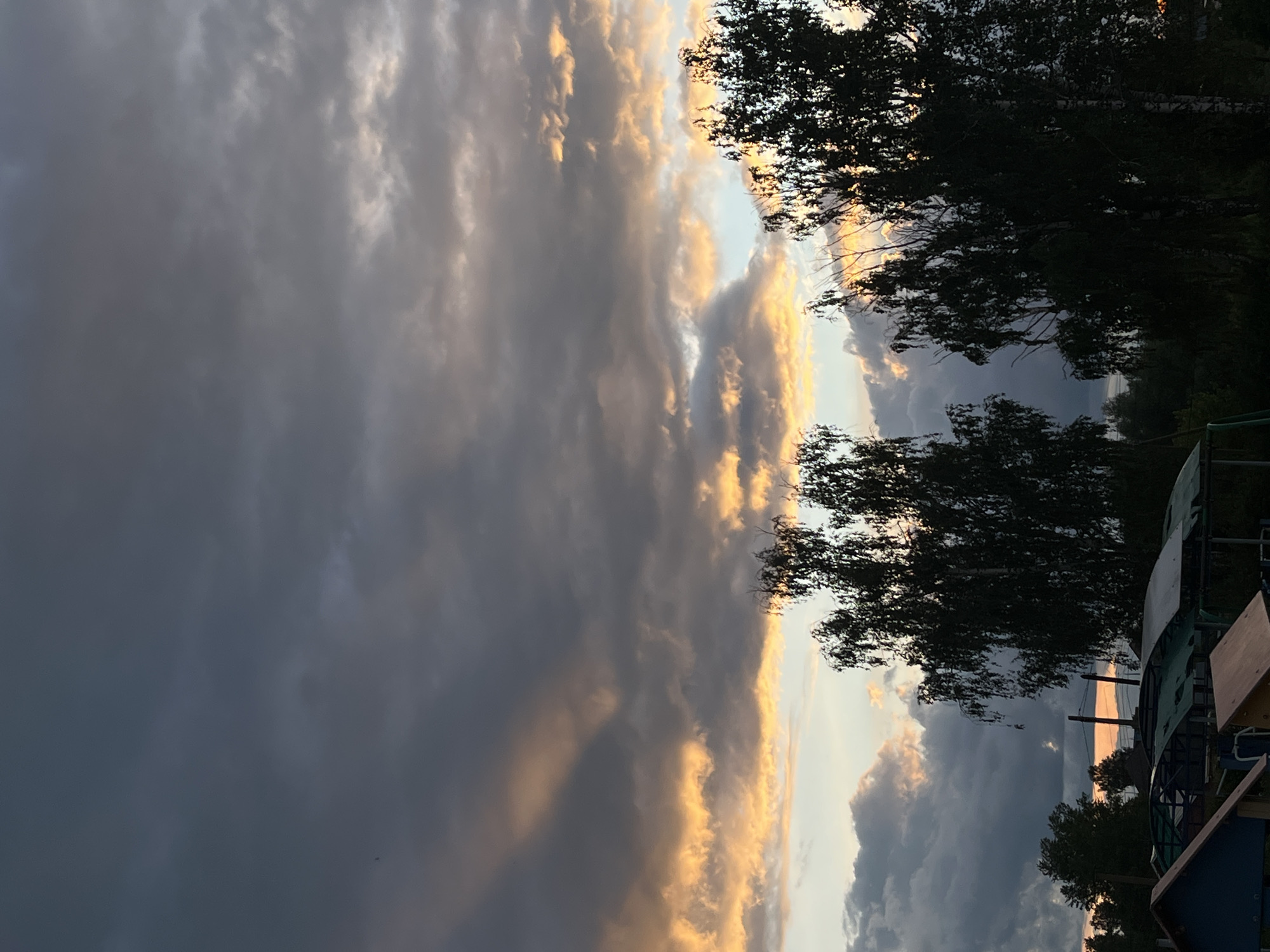 The image presents a view of the sky in Фоки, Russia. The sky is filled with clouds, illuminated by the setting sun, with the tree outlines in the foreground. Some man-made objects are partially visible at the lower right of the image.