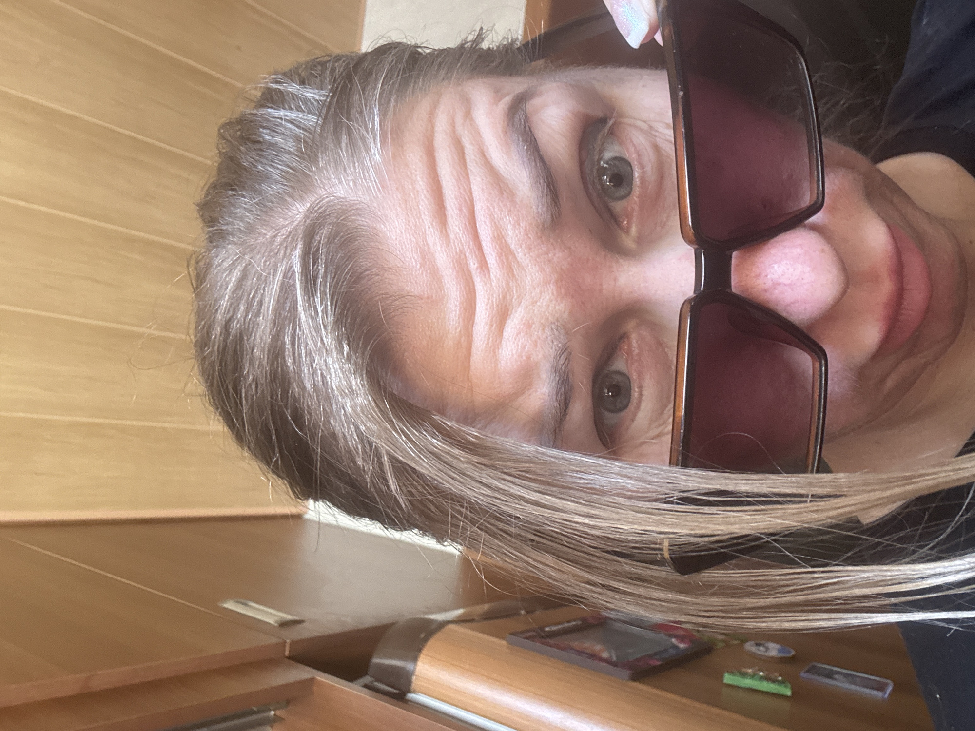 A close-up, slightly angled image depicts a person with graying hair, wearing sunglasses. The person has noticeable wrinkles on their forehead. The image captures the person's face, including their eyes, nose, and mouth. The background appears to show a wooden surface or piece of furniture. Several small objects are visible near the bottom of the image, on the same wooden surface as the furniture. The location is not discernible from the image.