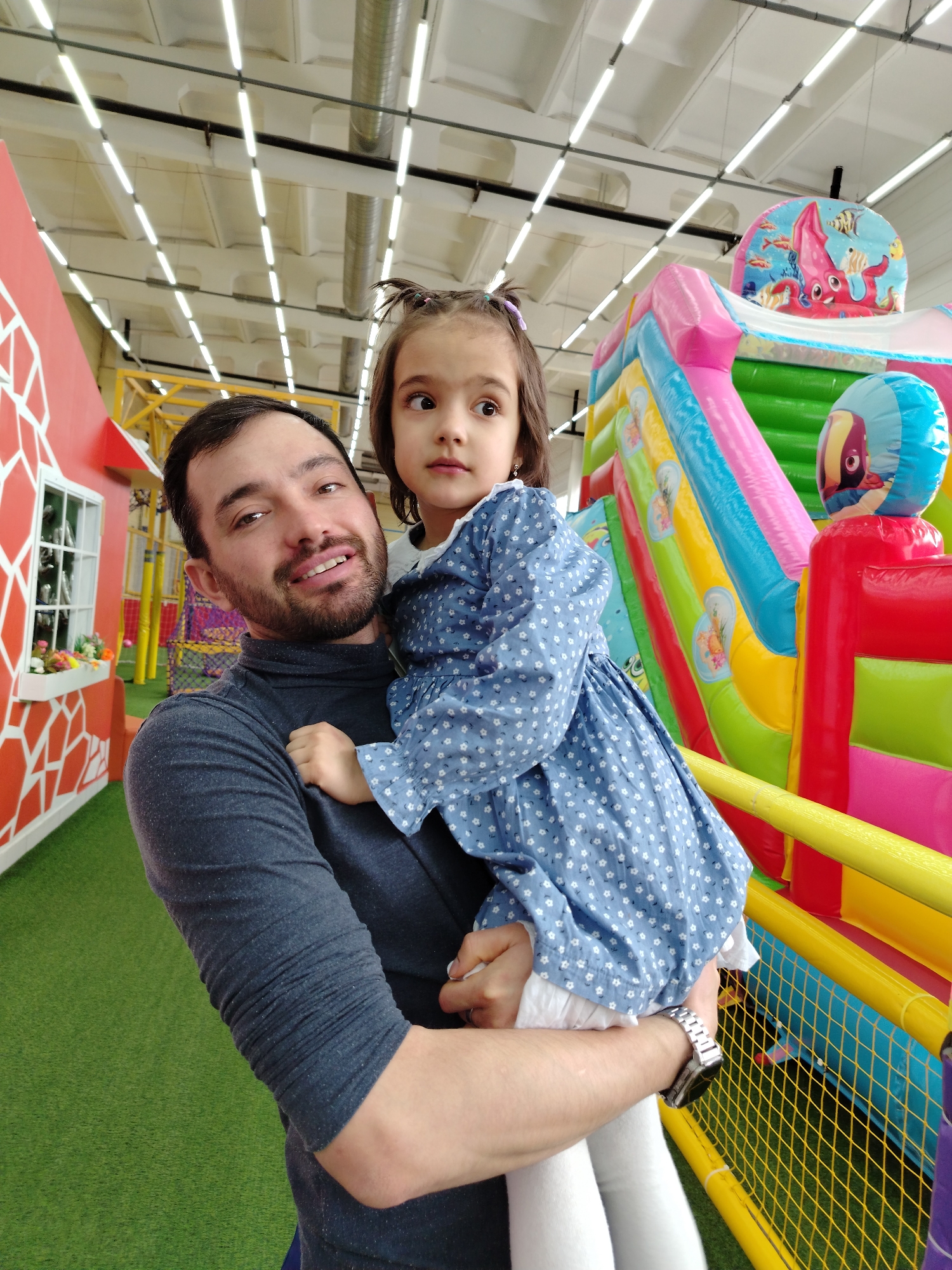 A man is holding a young girl in his arms inside a brightly lit indoor playground in the city of Unknown, Unknown. The man has dark hair and a beard, and he is wearing a dark grey long-sleeved shirt. He is looking at the camera with a slight smile. The girl, who is possibly his daughter, is wearing a blue dress with white flowers and white leggings. She has dark hair with a purple bow. The playground features a colorful inflatable slide with a sea-themed design, a red structure resembling a house, and artificial green grass. The ceiling is visible with fluorescent lights.