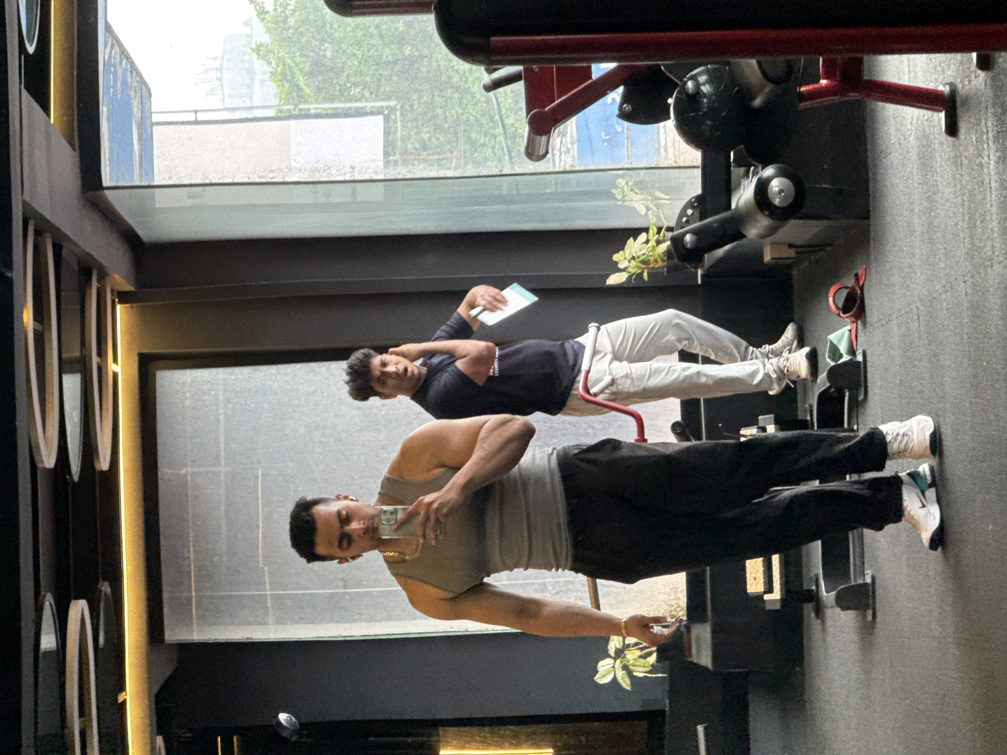 This image depicts an indoor scene in what appears to be a modern gym or fitness center, featuring two men.
In the foreground, a muscular man with dark hair, wearing a gray tank top, black pants, and white and teal sneakers, is taking a selfie using a smartphone. He has a gold chain around his neck and a gold bracelet on his left wrist. His reflection is clearly visible in a large mirror that forms part of the gym's wall.
Further into the background, a second man is positioned near a large window. He is wearing a dark blue t-shirt, light-colored pants, and light-colored sneakers. He appears to be resting or observing, holding a white rectangular object (possibly a notebook or tablet) in one hand, with the other hand near his head, while sitting on a piece of gym equipment. Parts of various gym machines, including one with red and black components, are visible around him.
The gym has a contemporary aesthetic with dark walls, dark flooring (possibly rubber gym mats), and some overhead lighting fixtures. A large window provides ample natural light, revealing an outdoor scene with green foliage and buildings, indicating it is daytime. A small potted plant is placed near the window, adding to the decor. No explicit text is visible within the image. The setting suggests an urban environment, and the provided context places this scene in Kolkata, India.