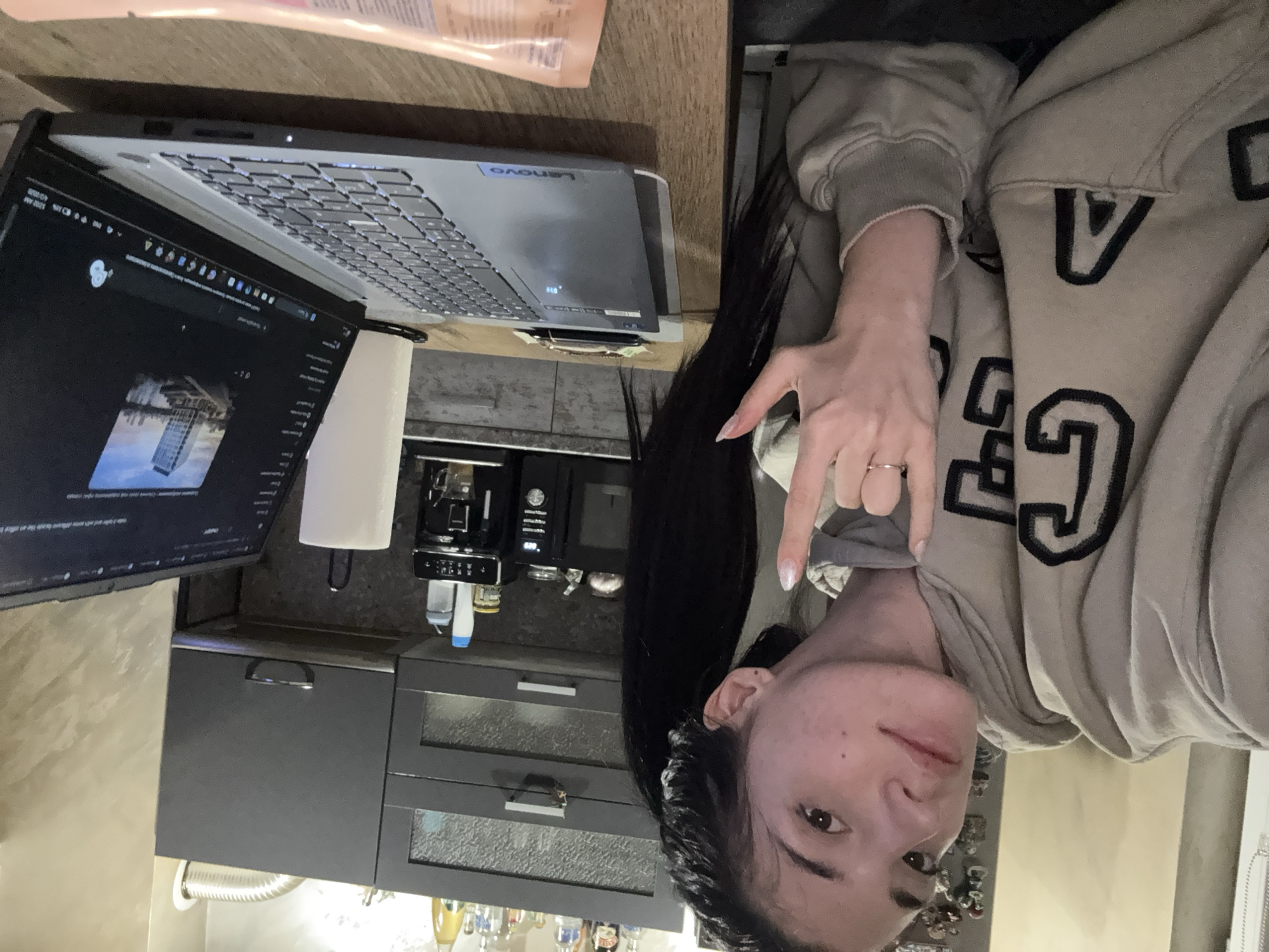 A woman wearing a gray hoodie makes a rock and roll gesture, sitting at a table with a Lenovo laptop and a second display screen. A tube of lotion rests to the left of the laptop. A coffee machine and kitchen cabinets are visible in the background. There is no indication in the image as to whether the location is in Sofia, Bulgaria.