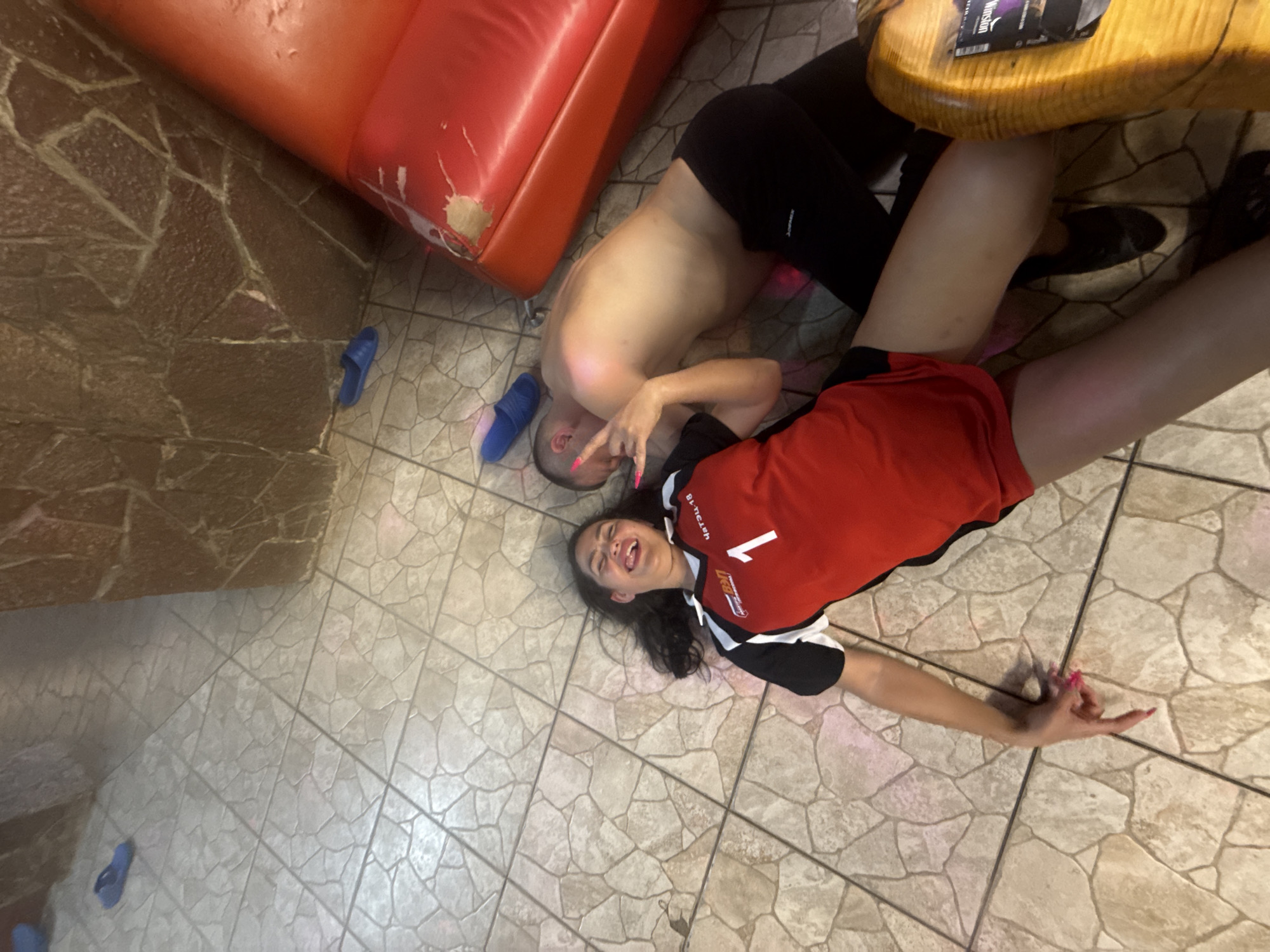 The image depicts a man and a woman lying on a tiled floor next to a red leather couch and a small wooden table. The man is shirtless and wearing black shorts, while the woman is wearing a red and black jersey. The woman is smiling and gesturing with her hand. The location is Chaikovsky, Russia, as indicated by the word "Чайковский" on the woman's jersey. Blue sandals are visible near the couch.