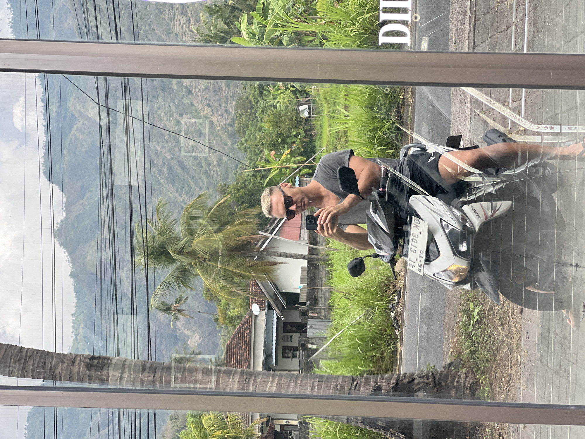 The image is a mirror reflection of a man on a white scooter in front of a rural landscape with mountains in the background. The man is wearing sunglasses, a grey t-shirt, and black shorts. He is holding a smartphone and taking a selfie. The scooter has a license plate that reads "DK 2033 KD". In the background, there are palm trees, houses, lush green vegetation, and power lines. The scene appears to be in Abang, Indonesia, based on the license plate format which is typical for Bali, Indonesia, and the tropical scenery. The reflection is captured in a large pane of glass, possibly a shop window, as indicated by a partial sign reading "DIL" on the right side of the frame.