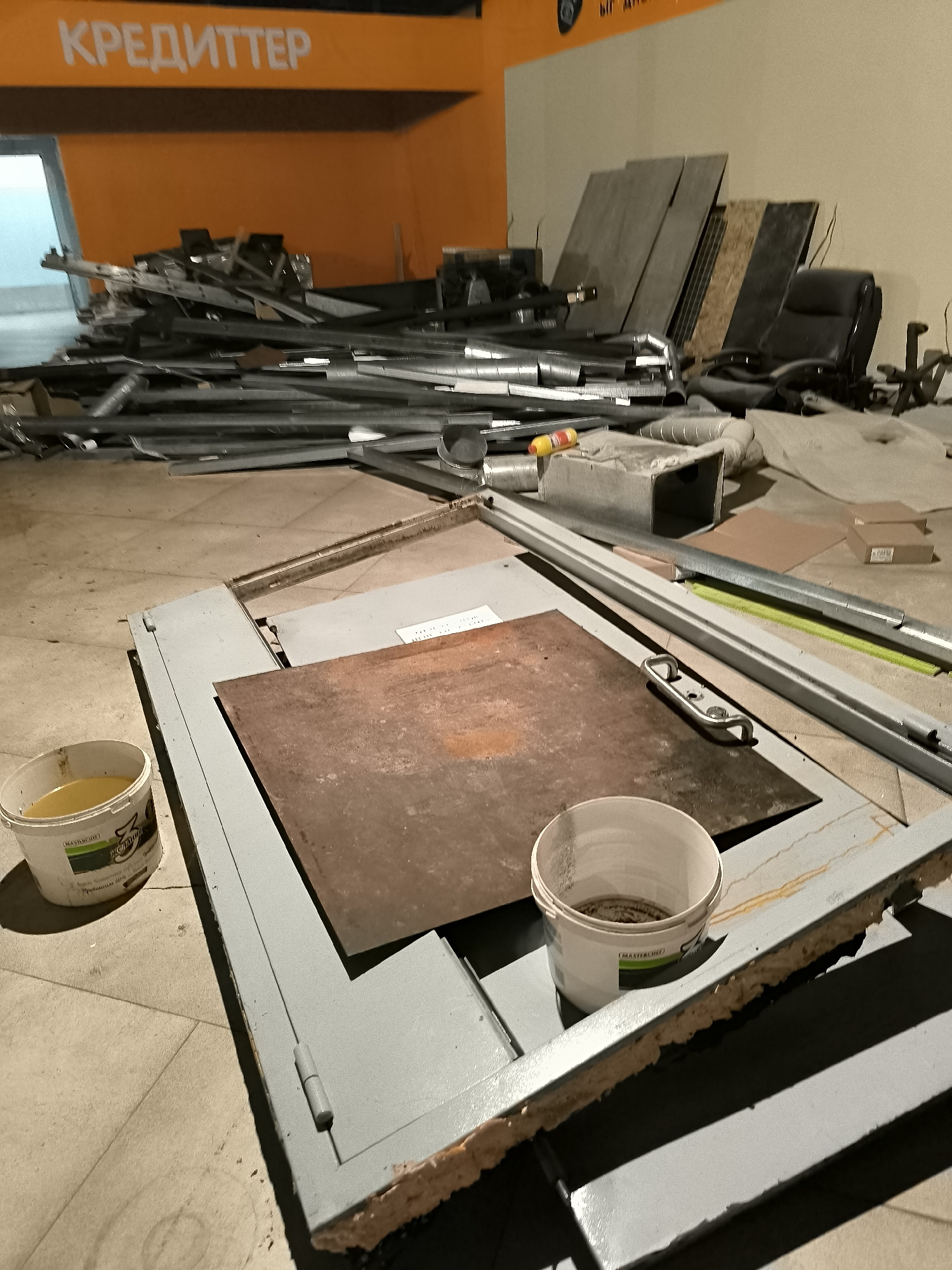 The image shows a construction or renovation site within an indoor space. In the background, an orange wall with the Cyrillic text "КРЕДИТТЕР" (which translates to "Creditter") is visible. A pile of metal debris, including pipes and framing, is scattered on the floor. A black office chair and several large, flat panels, likely construction materials, lean against the wall. In the foreground, a large, gray metal frame with hinges is laid flat on the floor. A rusty metal plate is placed on top of this frame. Two white buckets are present; one contains a yellow liquid, and the other is mostly empty with some residue at the bottom. The floor appears to be tiled or concrete. The overall impression is one of a disorganized workspace, possibly after a demolition or during a renovation phase. The location in Qarağandı city administration, Kazakhstan is not directly indicated by any visual cues in the image itself, but this context is provided.