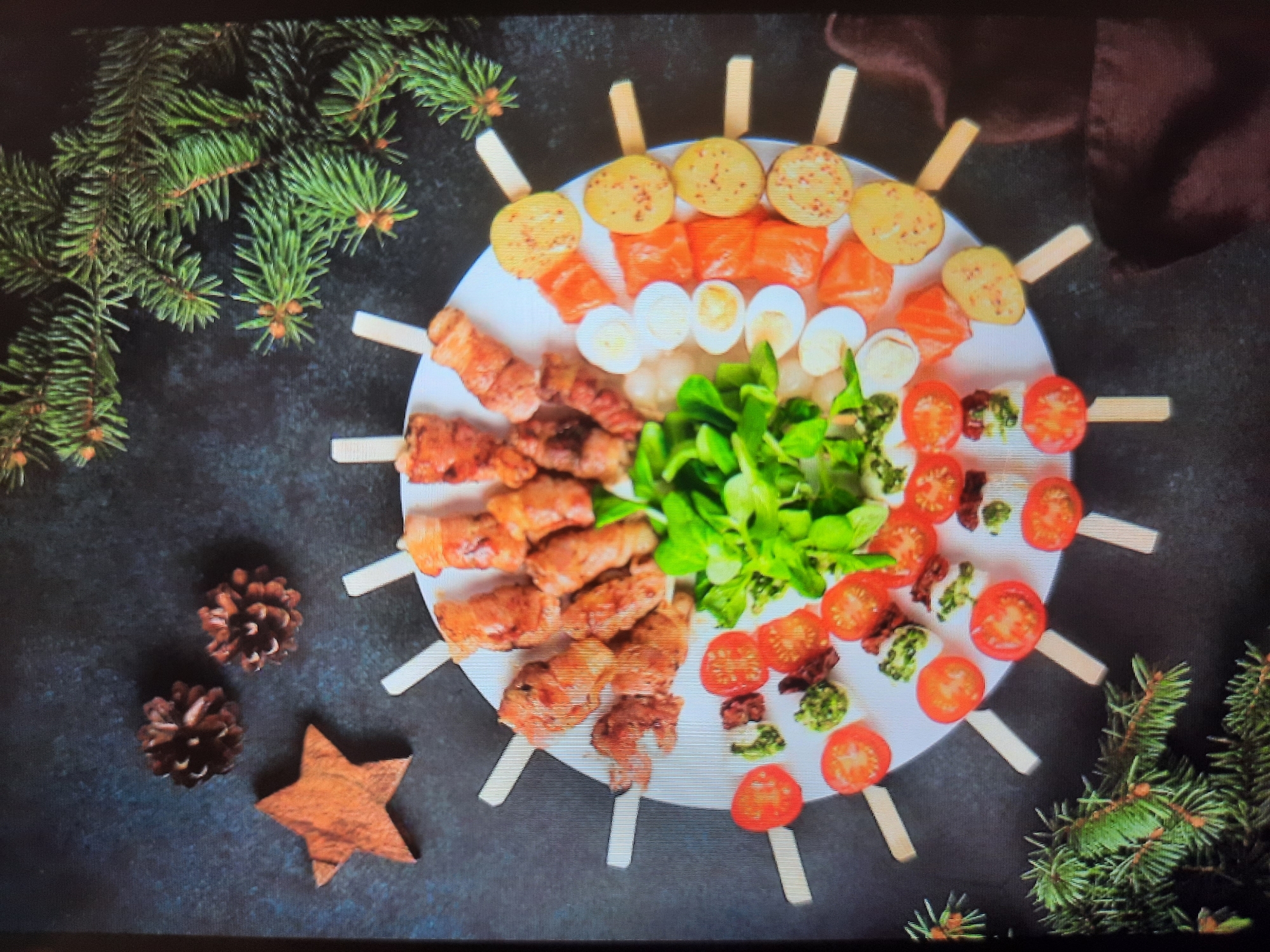 The image shows a circular platter arranged with food skewers, resembling a clock face. The skewers hold various items including meat wrapped in bacon, sliced tomatoes, quail eggs, pieces of salmon, and potato slices. In the center of the platter is a pile of fresh green leaves, likely a salad. The platter is placed on a dark, textured surface, possibly a table. Decorative elements like pine cones and a wooden star are visible in the upper left corner, and pine branches frame the platter from the top and bottom right. The lighting suggests an indoor setting, potentially a table set for a meal or a styled food photograph. There are no people present in the image. The location in the city of Rosenheim, Germany is not ascertainable from the image.