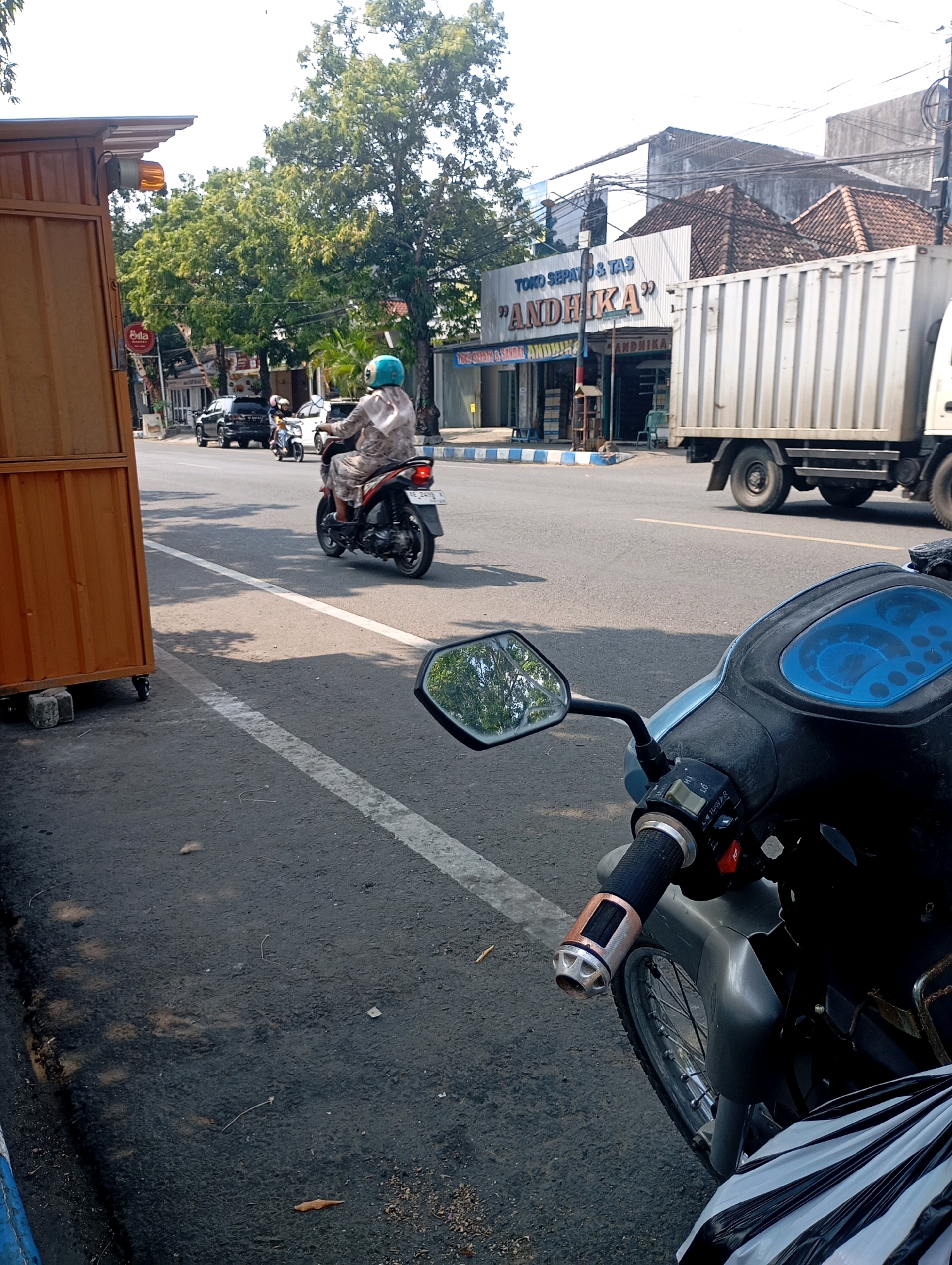 An eye-level shot captures a street scene in Pacitan, Indonesia, on a bright, sunny day. The foreground is dominated by the front of a parked motorcycle, with its handlebars and dashboard visible. To the left, a wooden cart, possibly for street vending, stands on the side of the road, with its orange hazard light illuminated.
The street itself is paved with asphalt and has a white dividing line. A person on a scooter, wearing a patterned outfit and a turquoise helmet, is riding away from the viewer towards the right side of the frame. Behind the scooter, other vehicles, including a black SUV and a white car, are visible.
Further down the street, buildings line the right side. One building has a sign that reads "TOKO SEPATU & TAS 'ANDHIKA'" (Shoes and Bags Store 'ANDHIKA'). A large white truck with a container is parked or moving slowly on the right side of the road, partially obscuring the buildings. Trees with lush green foliage are interspersed along the roadside and behind the buildings, providing shade. The sky is clear and bright. The overall impression is of a typical urban street environment in Indonesia during daytime.