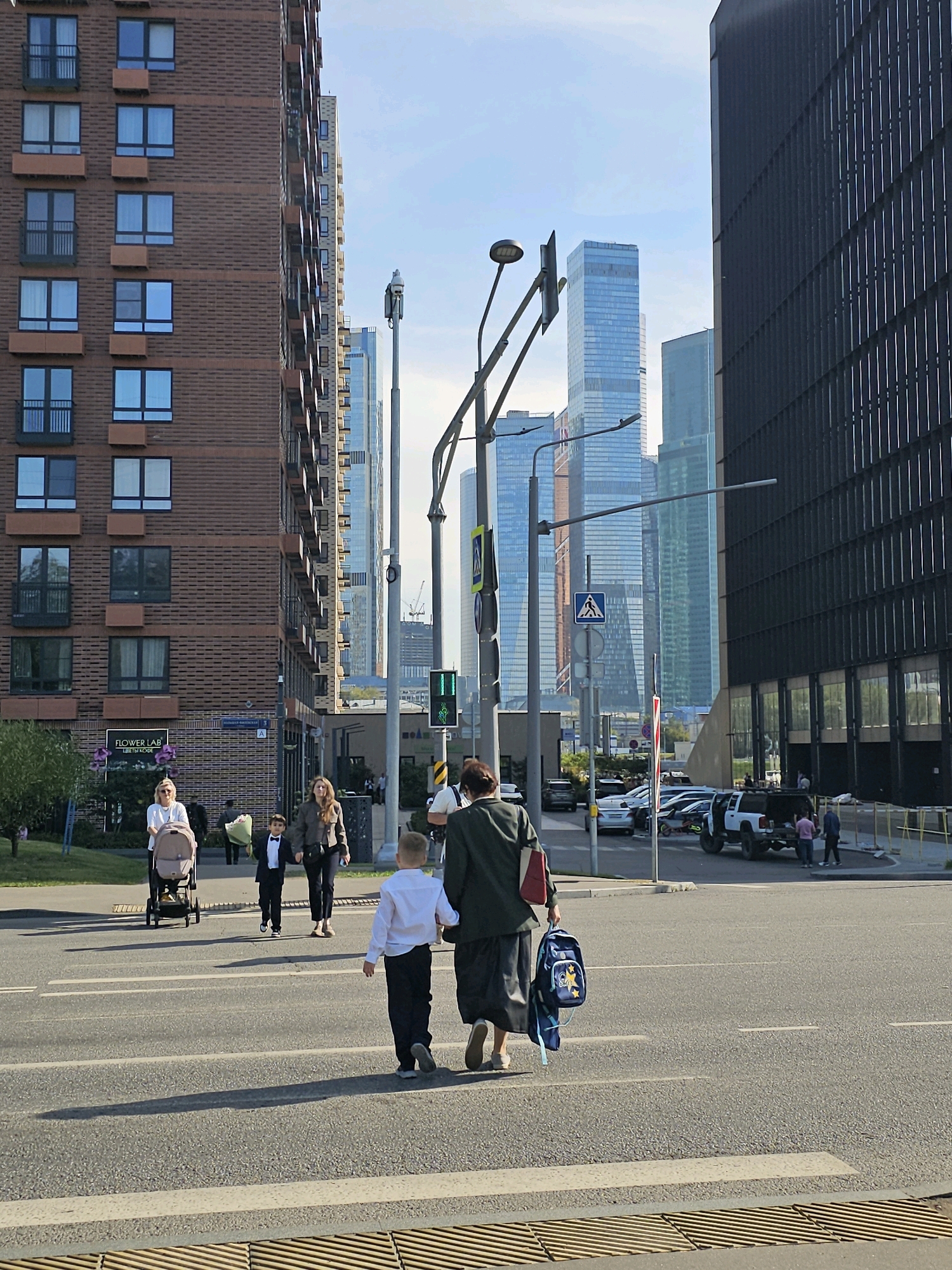 A street scene in Moscow, Russia, shows a group of people crossing a road. The foreground features a pedestrian crossing with a tactile paving strip. On the left, a woman pushes a stroller with a child inside. Beside her, a young boy in a formal suit and bow tie walks with a bouquet of flowers. Further ahead, a woman walks with a blue backpack.
In the background, several modern skyscrapers dominate the skyline, including the prominent Moscow City business district. Buildings on the right are dark and imposing, with vertical window patterns. On the left, a brick residential building with many windows is visible. Traffic lights, street lamps, and street signs are present. Cars are parked and moving on the road and sidewalks. The sky is mostly clear with a few scattered clouds.