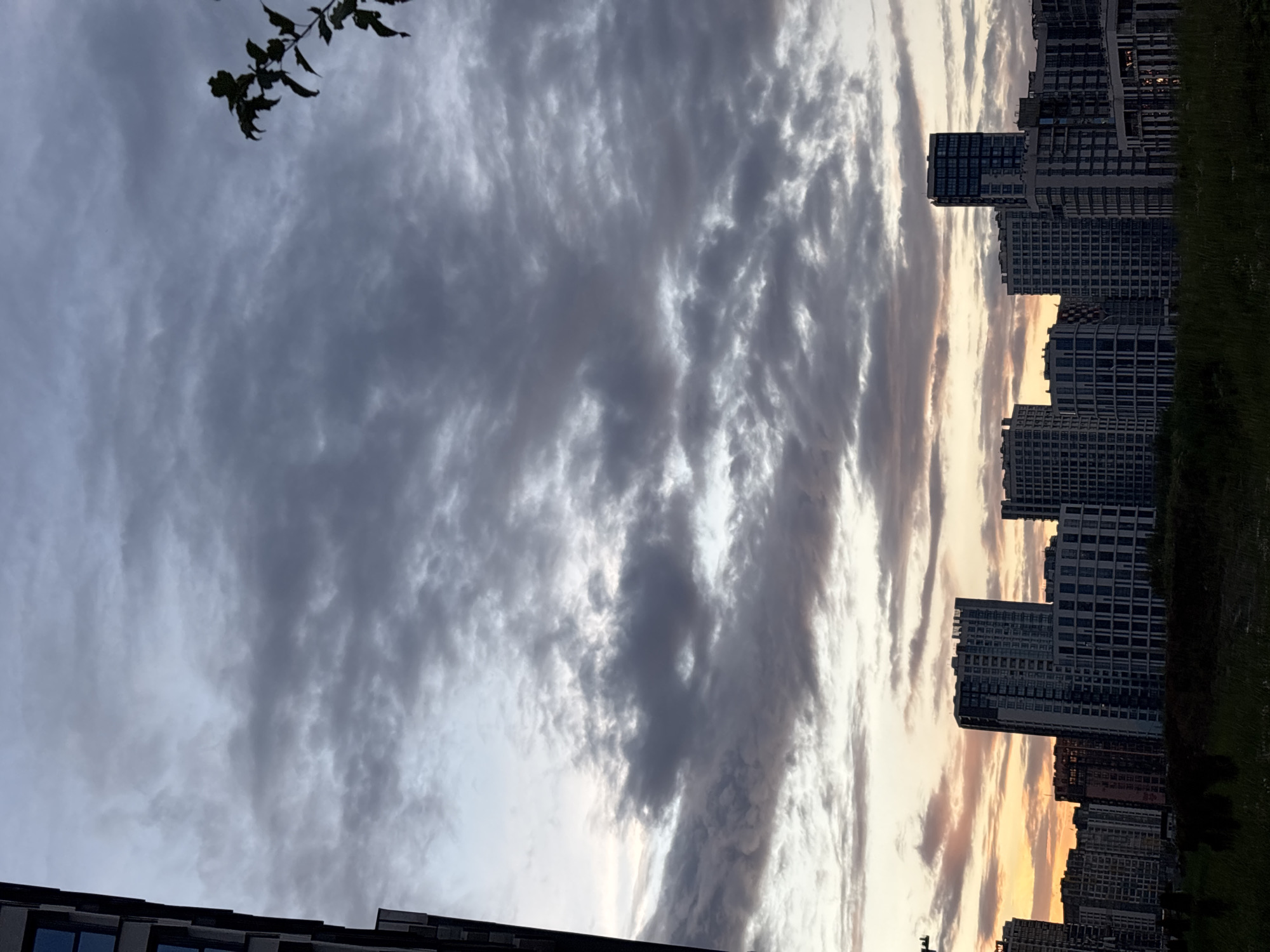 The image depicts a skyline of modern buildings at dusk in Minsk, Belarus. A large, dark building occupies the left edge of the frame, partially obscuring the sky. The sky is filled with dramatic, cloudy formations, ranging in color from deep gray to lighter tones, with hints of orange and yellow near the horizon, suggesting the sun is setting. A silhouette of tree branches is visible in the upper right corner. Below the sky, a row of multi-story residential buildings stretches across the frame. These buildings vary in height and architectural style, with many featuring numerous windows. In the foreground, a dark, silhouetted area suggests dense grass or vegetation. The scene is captured in a way that emphasizes the imposing presence of the architecture against the atmospheric sky. There are no visible people or ongoing actions.