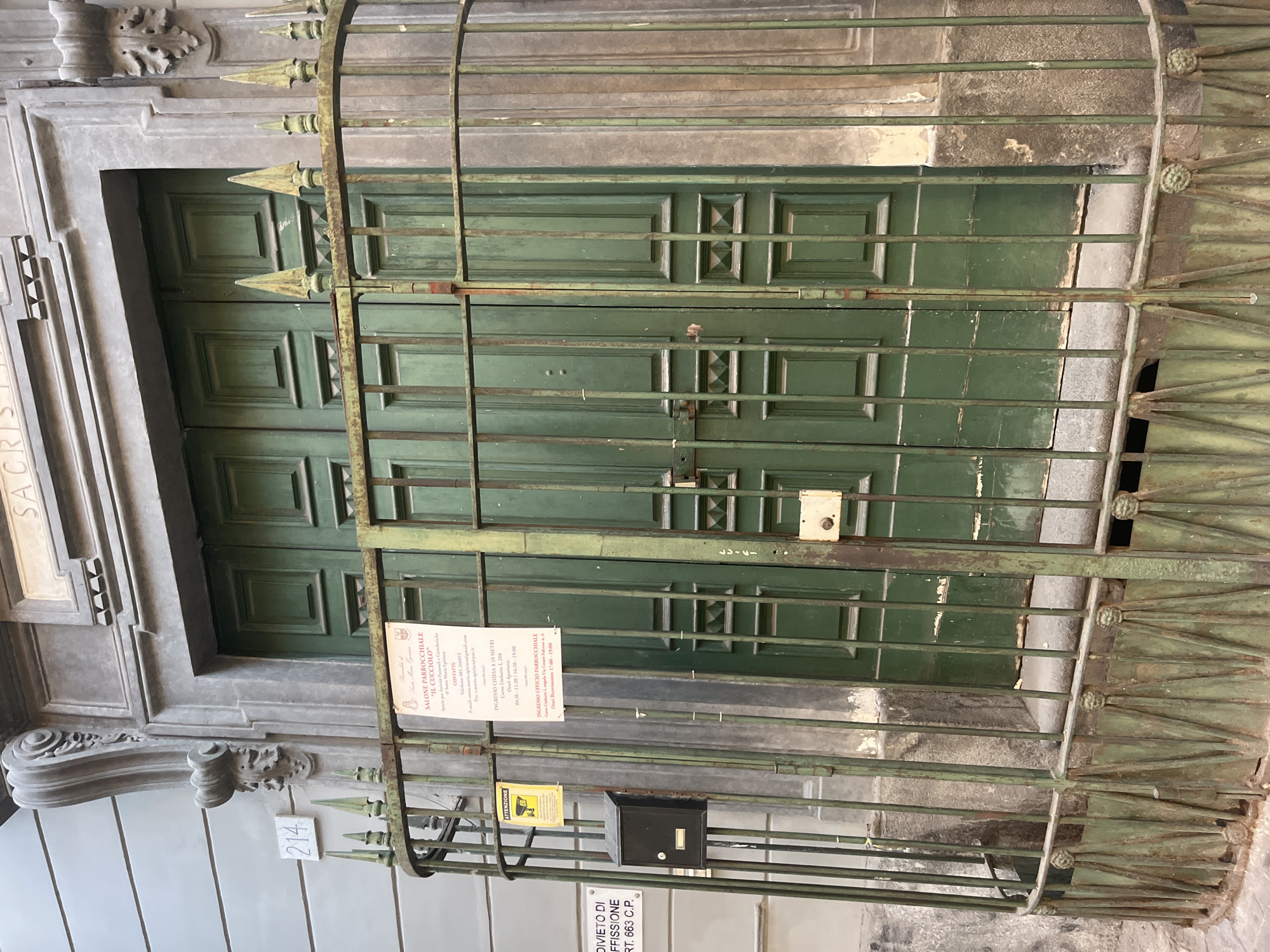 The image shows a weathered green double door behind a green metal gate in Naples, Italy. A sign with Italian text is affixed to the gate. Above the door, the word "SACRISTIA" is visible on the building's facade. A street number "214" is also visible on the building's left side. The scene appears to be an entrance to a church or religious building.