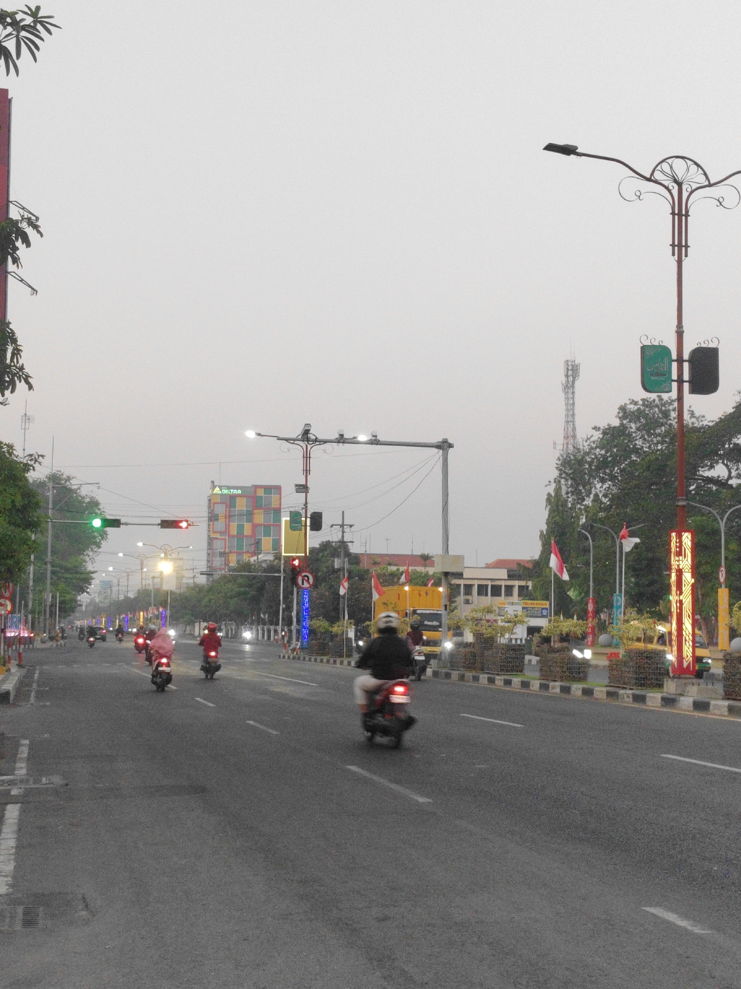 The image shows a street scene in Sidoarjo, Indonesia, during daylight hours with overcast skies. The perspective is from ground level, looking down a multi-lane road lined with trees, streetlights, and buildings. Several motorcycles are visible on the road, with riders wearing helmets and casual clothing. One motorcycle in the foreground has its brake light illuminated. Traffic signals are present at an intersection, showing green and red lights. A tall communication tower stands in the distance. Buildings on either side of the street vary in architectural style, with one prominent building featuring a colorful, multi-colored facade. Indonesian flags are displayed on poles along the roadside. The overall impression is of a moderately busy urban environment.