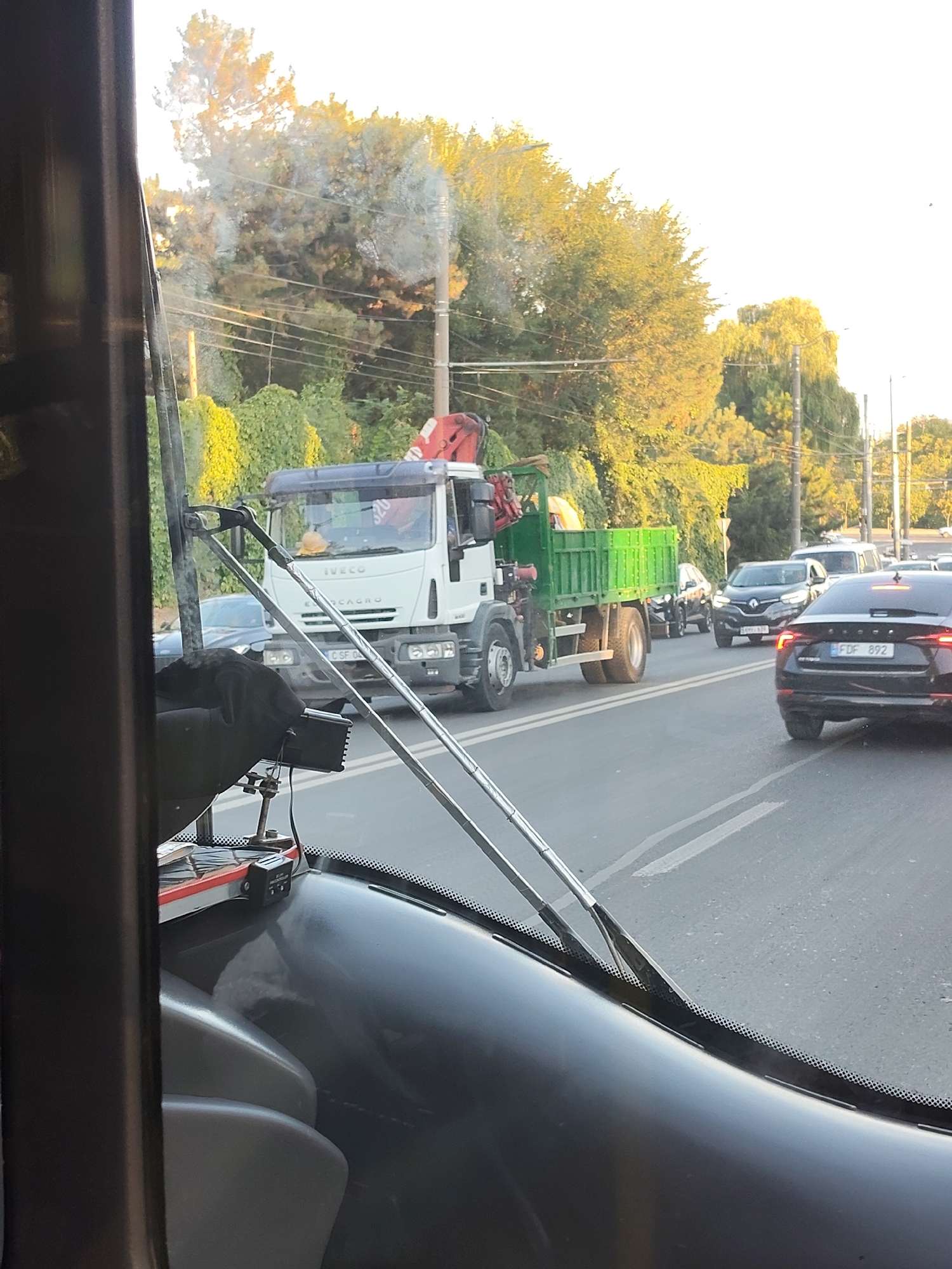 The image shows a view from inside a vehicle, looking out through the windshield. It appears to be daytime with bright sunlight. The vehicle is on a road, likely in Chișinău, Moldova, given the context.
In the foreground, parts of the vehicle's interior are visible, including the dashboard, seats, and windshield wipers. The dashboard is dark and appears to be made of plastic. The windshield is reflective, showing a hazy view of the outside.
Outside the vehicle, there is traffic on a multi-lane road. The most prominent vehicle is a white Iveco truck with a green flatbed carrying what appears to be construction debris or materials. The truck has a crane attached to its rear. A yellow hard hat is visible on the dashboard of the truck.
Other vehicles in traffic include a dark-colored SUV or sedan with a license plate that reads "FDF 892" and a black Renault sedan. There are also other cars and possibly a white van further down the road.
The environment includes trees and lush green vegetation lining the side of the road. There are utility poles with wires visible against the bright sky. The overall impression is of a busy street scene in an urban or suburban area. The light suggests it might be late afternoon or early evening, with the sun casting a warm glow on the trees.