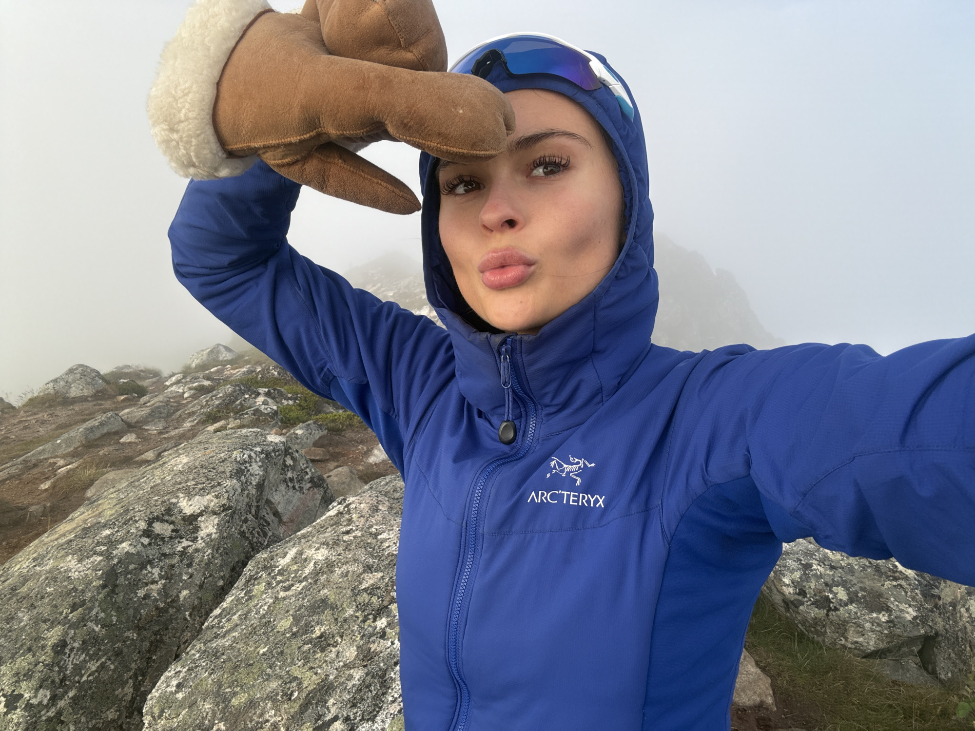 A woman is taking a selfie while wearing a blue hooded jacket with the Arc'teryx logo, goggles, and a brown glove on her left hand, which is raised to her forehead. She is making a kissing face. She is standing on a rocky, mountainous terrain with fog in the background. The location is specified as Harstad, Norway.