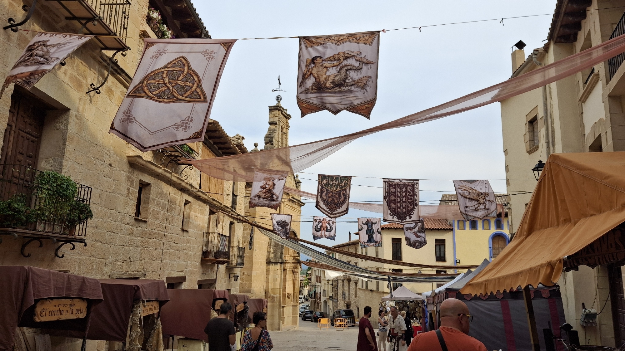 The image depicts a street scene in the city of La Fresneda, Spain, likely during a medieval fair or festival. Banners with various historical or mythical illustrations and crests are suspended across the street, adding a festive and thematic atmosphere. The architecture features stone buildings with wooden balconies and shuttered windows, characteristic of traditional Spanish towns. Several market stalls are set up along the street, covered with brown awnings, suggesting a commercial or artisanal event. People are visible browsing the stalls and walking along the street, some in modern attire while others appear to be in costume, further hinting at a historical reenactment or themed celebration. The sky is overcast, casting a soft light over the scene. The overall impression is one of a lively cultural event taking place in a historically preserved urban setting.