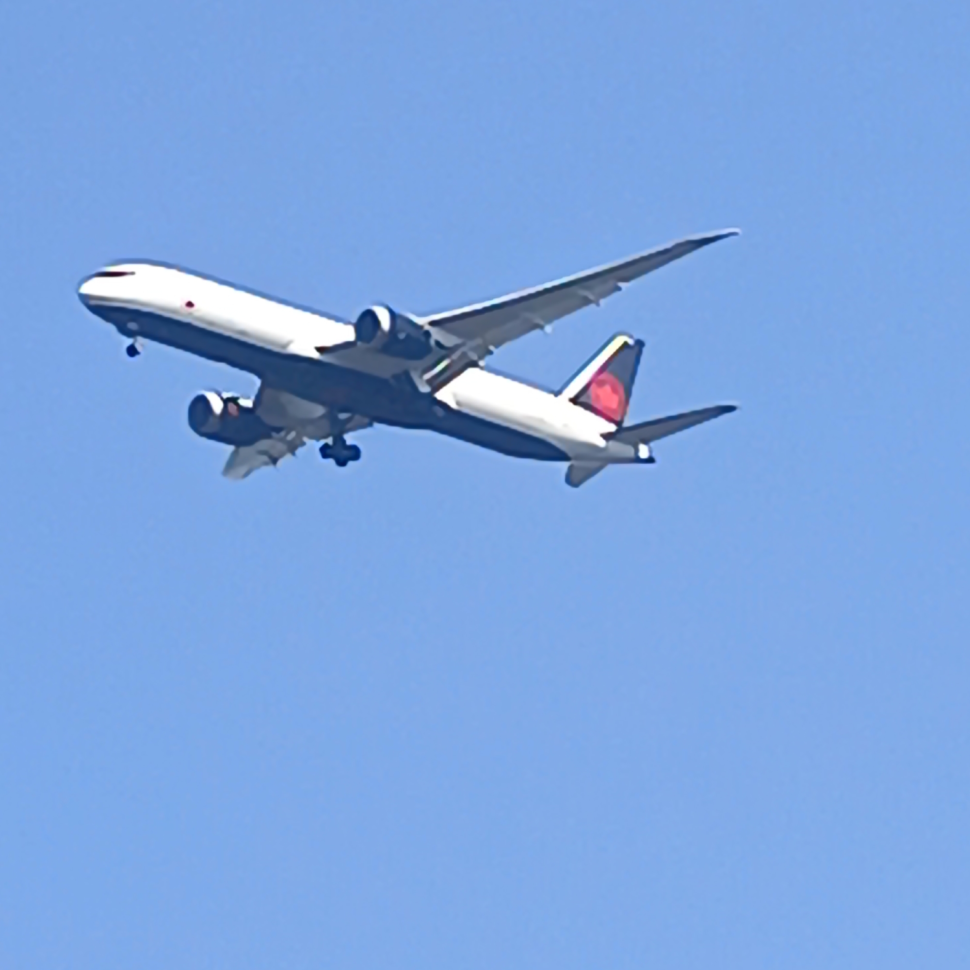 A wide-body jet aircraft with white fuselage and dark blue accents on the wings and tail is captured in flight against a clear blue sky. The aircraft's landing gear is extended. The tail fin features a red triangular logo. The image does not provide any information about the city of Frankfurt, Germany, or any specific location. There are no people or interactions visible.