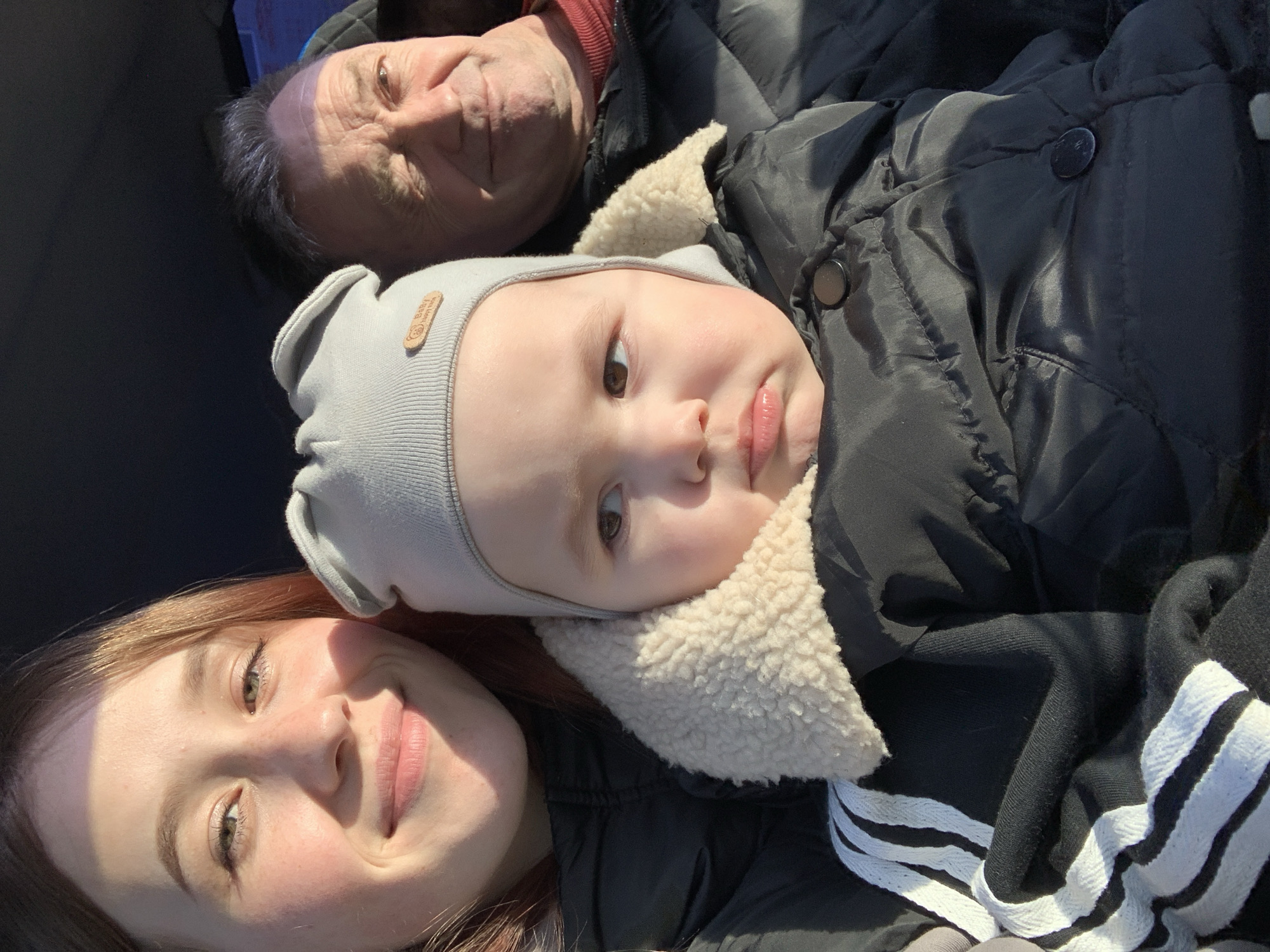 A close-up, diagonal portrait captures three individuals, likely spanning three generations: an older adult male, a baby, and a young adult female, positioned in a tight group, possibly for a selfie. The image is taken in bright daylight, casting shadows on their faces.
At the top, an adult male with dark hair (showing a faint purple hue, possibly from light or hair dye) and a light smile looks towards the camera. He wears a dark jacket over a reddish collar. Below him, in the center, a baby gazes directly at the camera with a neutral, slightly curious expression. The baby wears a light grey ribbed beanie adorned with a small wooden tag that reads "Baby Happy Day" and is bundled in a black puffy jacket featuring a cream-colored, fluffy collar. At the bottom, a young adult female with light brown hair smiles warmly towards the viewer. She is wearing a dark top, with a visible sleeve showing black fabric with three white stripes, characteristic of an Adidas garment.
The subjects appear to be outdoors or in a vehicle, given the strong, natural lighting and indistinct, dark background. Their warm clothing suggests cool weather. The overall mood is affectionate and pleasant. The location context provided is Фоки, Russia.