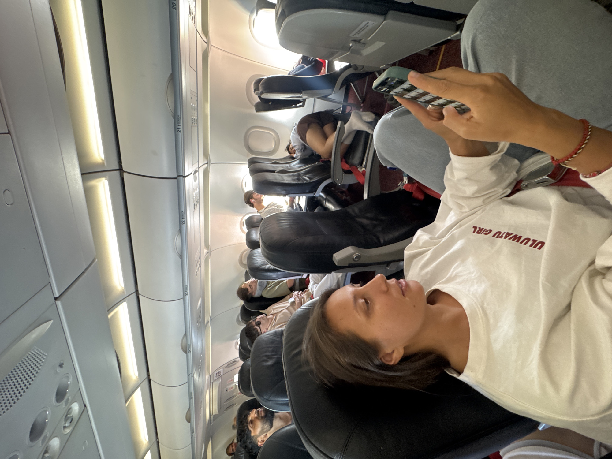 The image displays the interior of a commercial aircraft cabin, observed during what is stated to be a journey to or from Đà Nẵng, Vietnam. In the immediate foreground, a light-skinned female with medium-length dark hair is seated in a black airline seat. She wears a white long-sleeved shirt with "ULUWATU GIRL" printed in red on the right chest area and a red, white, and gold braided bracelet on her left wrist. She holds a light-green cased smartphone with both hands, manipulating the screen with her thumbs, and her gaze is directed towards the device. Her lower body is clad in light blue jeans.
Behind the foreground subject, multiple passengers occupy black airline seats. To her direct left, a male with dark hair and a beard is visible, looking towards the left. Further down the left aisle, another male passenger is seen wearing sunglasses. On the right side of the aircraft, across the aisle, a passenger wears a grey neck pillow and appears to be sleeping, slumped in their seat. Behind this individual, a person occupies a window seat, curled up on their side with knees drawn up, wearing dark shorts and white socks, also appearing to be asleep or resting. Further back on the left, a passenger can be seen using a laptop.
The cabin environment features light-grey overhead bins with visible seat row numbers (e.g., 20, 21, 22), white rectangular overhead lighting fixtures, and oval-shaped windows emitting bright daylight. Red seatbelts are visible on several seats, and the aisle and under-seat areas show red floor covering. A partial "EXIT" sign is visible above a door-like structure in the background on the left side of the aisle. The image itself contains no explicit visual indicators such as airline logos or external landmarks to specifically confirm the flight's exact location within Đà Nẵng, Vietnam.