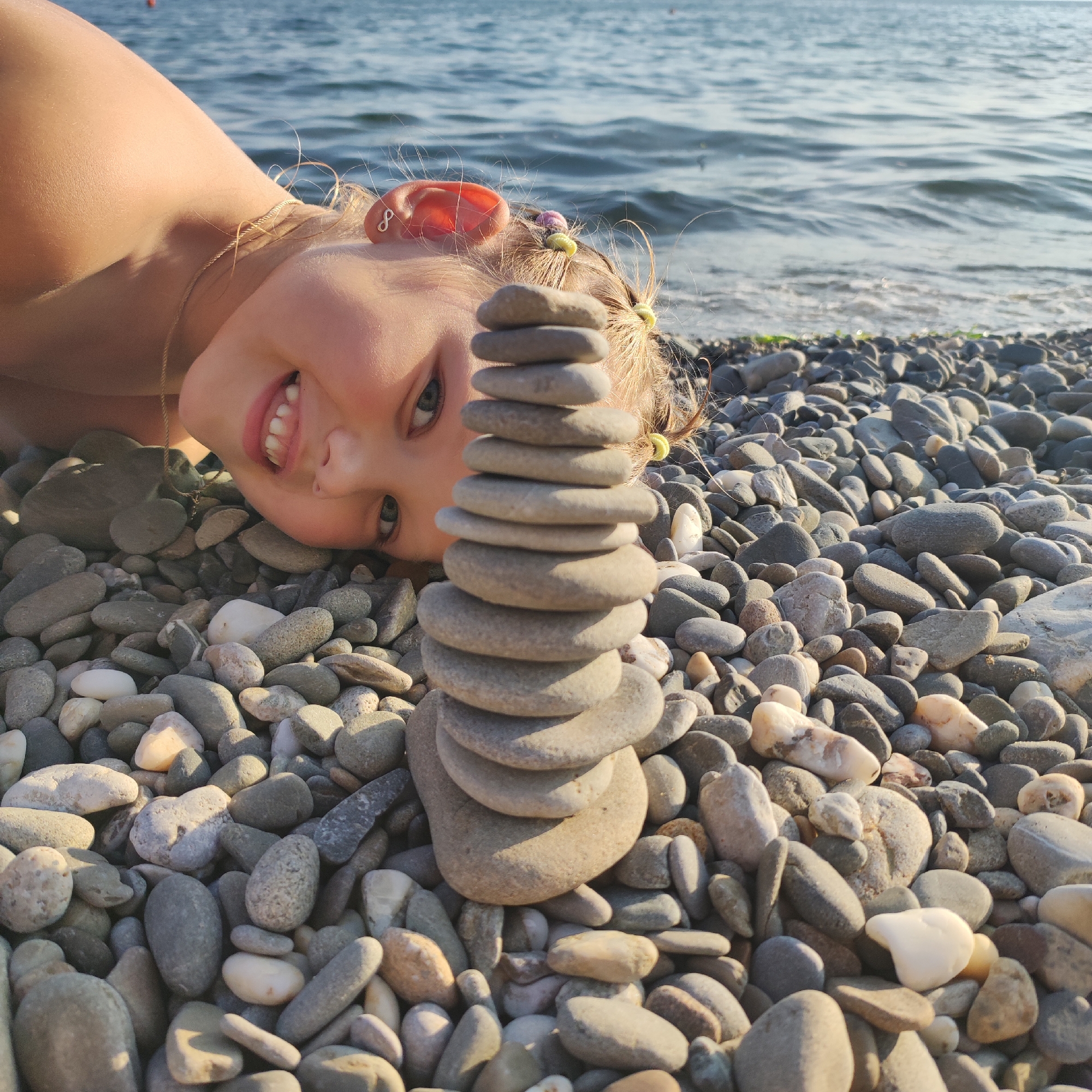 A young girl with blonde hair, wearing a tan top, lies on a rocky beach with her head tilted upside down and her face looking towards the camera. She is smiling, showing her teeth. Her hair is adorned with colorful beads and a small infinity symbol earring is visible on her left ear. In front of her face, a stack of smooth, grey pebbles has been artfully arranged. The ocean waves are visible in the background, with the water appearing blue and slightly choppy. The beach is covered in a dense layer of grey and tan-colored pebbles of various sizes. The sun casts a warm light on the scene, creating shadows on the pebbles and highlighting the girl's skin.