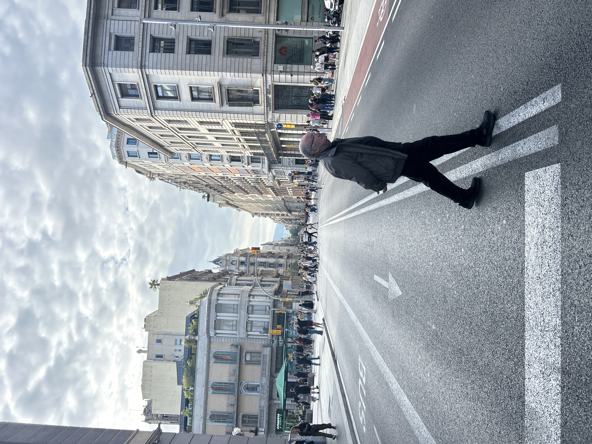 The image captures a street scene in Barcelona, Spain, viewed from a slightly tilted perspective. A man is positioned diagonally across a white crosswalk marking on the road, appearing to be either lying down or performing a similar action. He is wearing a dark jacket, dark pants, and black shoes.
The background showcases the urban architecture of Barcelona, with buildings lining the street. Some of the buildings have multiple stories and feature balconies or architectural details. A group of people is standing on the sidewalk. In the distance, the street continues, and more buildings can be seen. The sky is cloudy. The roadway is marked with a white arrow.