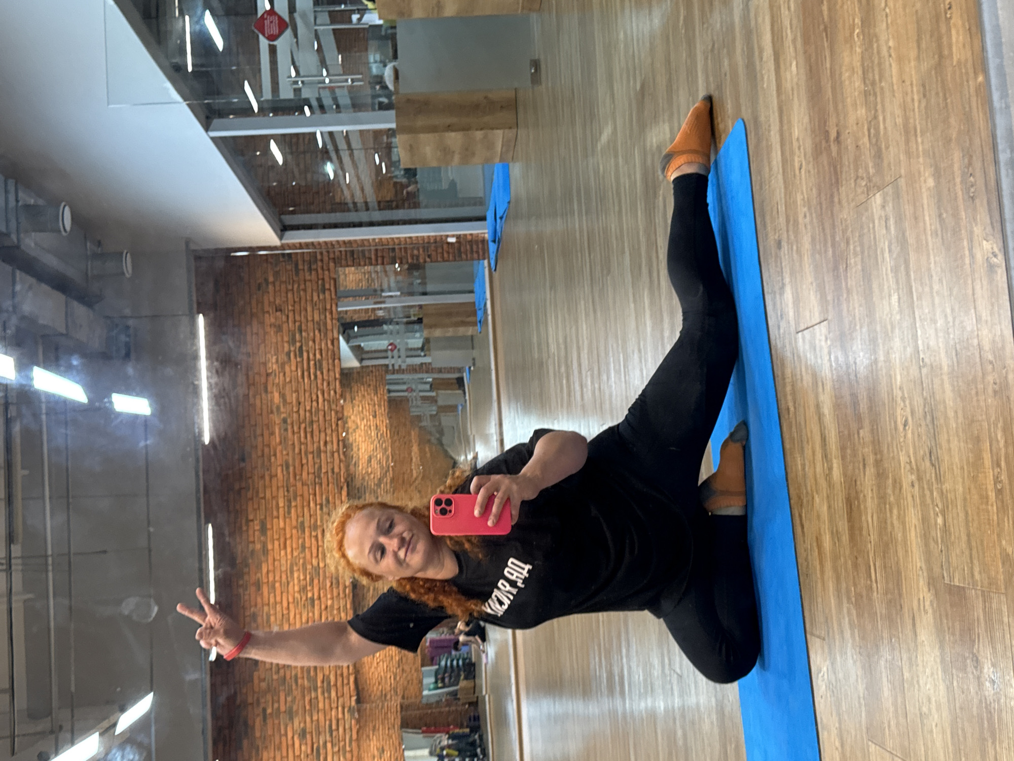 A woman with curly red hair, wearing a black t-shirt and black leggings with orange socks, is taking a selfie in a gym. She is kneeling on a blue yoga mat, with her left leg bent and her right leg extended forward. Her left arm is raised, with her hand making a peace sign. She is holding a pink smartphone in her right hand, with which she is taking the photo. The gym has a wooden floor, brick walls, and large glass windows. There are mirrors on the walls, and gym equipment can be seen in the background. The scene suggests a fitness or yoga studio. The location is not specified as Chaikovsky, Russia.