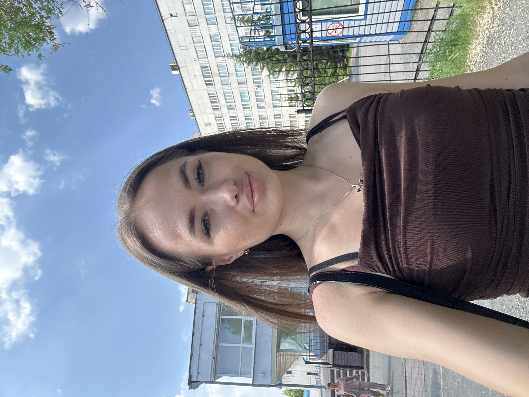 A young woman with long, brown hair and light skin is taking a selfie outdoors. She is wearing a brown, ruched tank top and a silver necklace with a pendant. Her head is tilted slightly to the side, and she is looking at the camera with a neutral expression.
Behind her, a light-colored building with many windows is visible. To the left of the building, there is a smaller structure with large windows and a covered walkway. In the background, there is a black metal fence and a blue and white structure with a "no smoking" sign. There are trees and green grass around the fence.
The sky is blue with scattered white clouds. The lighting suggests it is a sunny day. A person in a beige tracksuit is walking away from the camera on a paved path to the left. The ground is a mix of pavement and gravel.