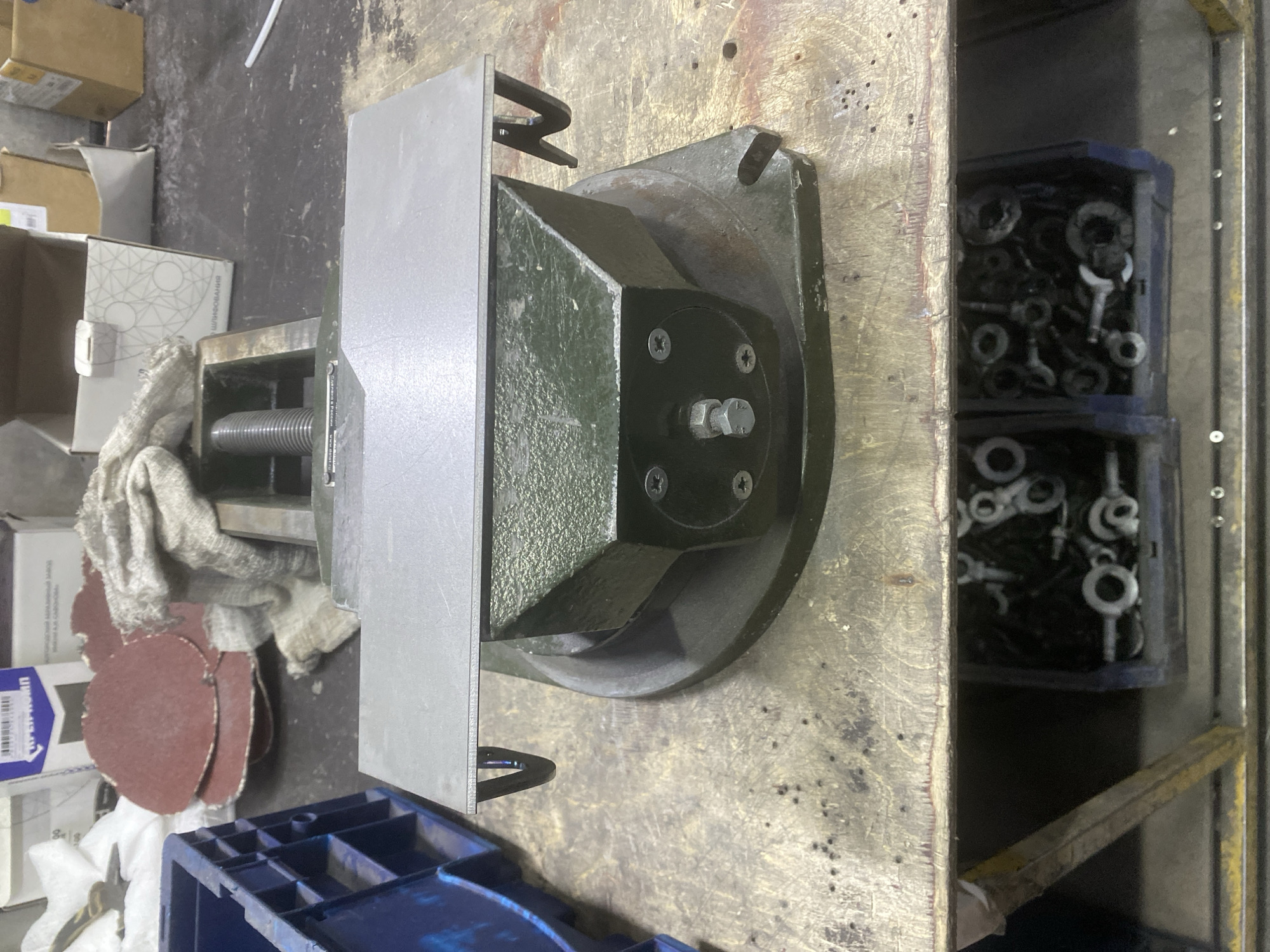 The image shows a workbench with various objects on it. The main subject is a large, dark green metal vise with a silver metal plate attached to its jaw. The vise appears to be stationary, and no activity is taking place. To the left of the vise, there are several round, red sanding discs, a white cloth, and some cardboard boxes. To the right of the vise, there are two blue plastic containers filled with various metal fasteners, such as nuts and washers. The workbench surface is rough and appears to be made of wood or metal, with visible wear and tear. The background is out of focus, but it seems to be an indoor setting, possibly a workshop or garage. There is some visible text on a blue and white box near the sanding discs, but it is difficult to read. The lighting suggests it is daytime. The location context of Berezhki, Russia, provides a general geographical setting.