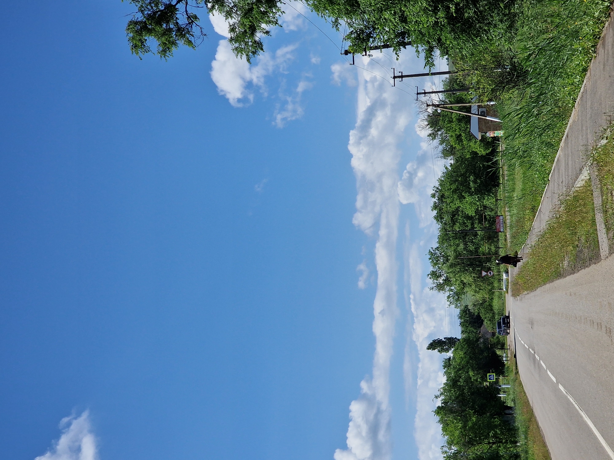 The image depicts a rural road scene in Kursavskaya, Russia. The road, paved with asphalt and marked with a white dashed line, curves slightly to the left and recedes into the distance. On the right side of the road is a concrete sidewalk bordered by tall, green grass and vegetation. A person, dressed in dark clothing and a headscarf, walks along the sidewalk away from the viewer. Further down the road, a dark-colored SUV is seen driving towards the viewer. Trees line both sides of the road, with dense green foliage. In the background, there are more trees, a few small buildings, and utility poles with electrical wires. The sky is a clear blue, with scattered white cumulus clouds. The overall impression is a quiet, sunny day in a rural setting.