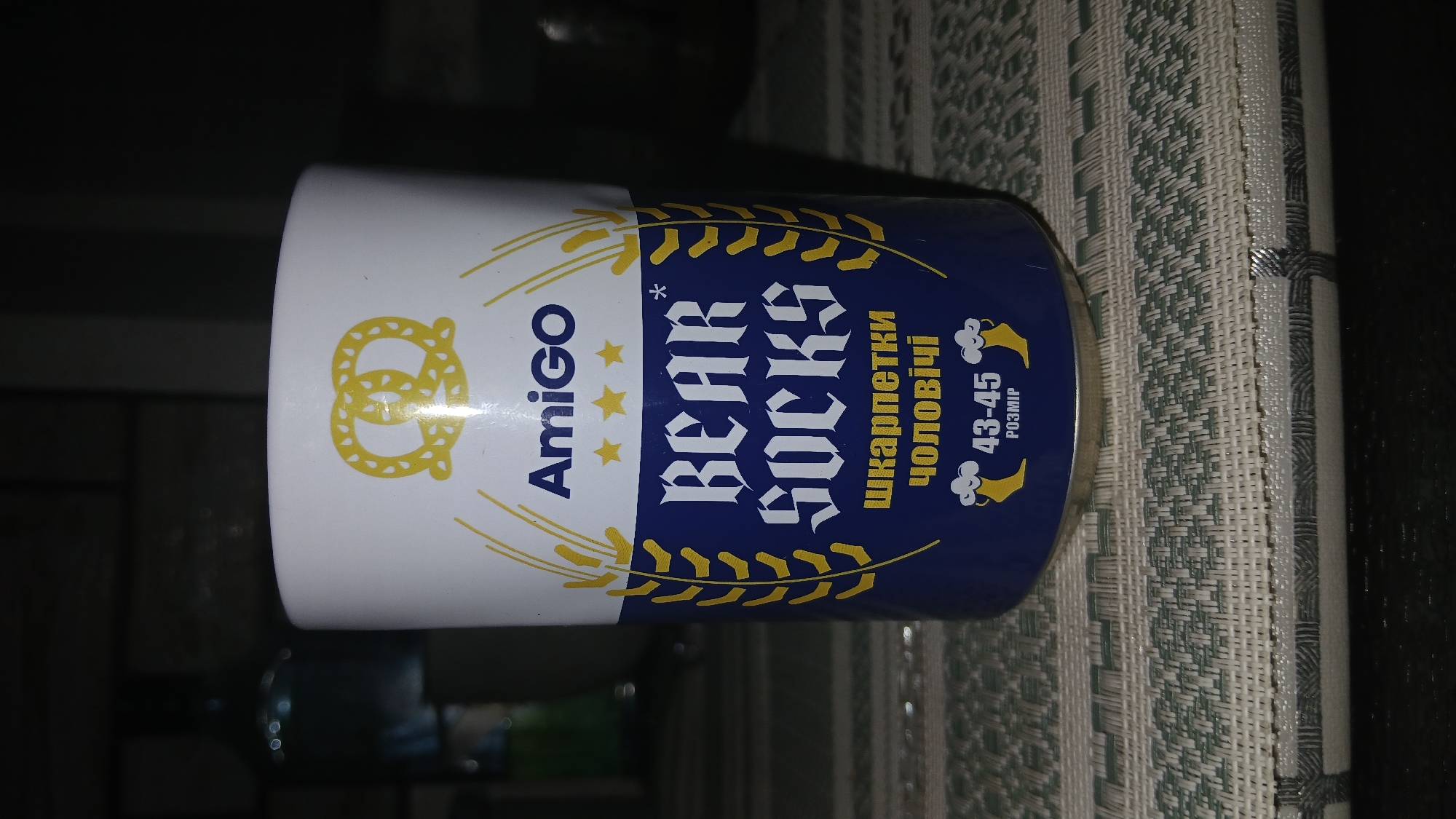 A cylindrical container with the branding "Amigo BEAR SOCKS" sits on a patterned surface. The container has a white top section adorned with a pretzel graphic. The lower section is dark blue and features the text "BEAR SOCKS" in stylized font, along with Ukrainian text. The size is indicated as "43-45" with a small graphic of feet inside the container. The location is Dnipro, Ukraine, based on the Ukrainian text on the container.