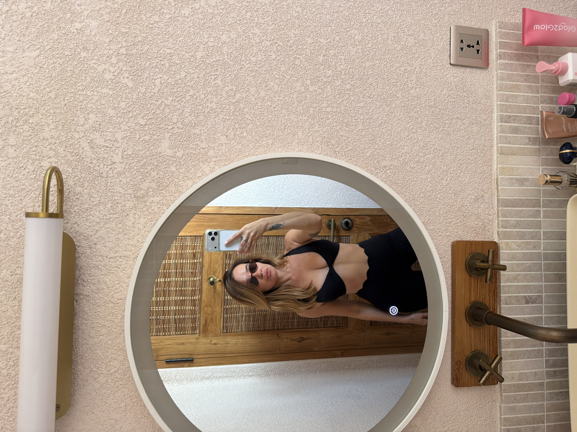 A woman is taking a mirror selfie in a bathroom. She is wearing a black bikini top and black bottoms with a scalloped edge. She has on sunglasses and her hair is down. The mirror is round with a light-up edge. Behind her, a wooden door with woven details is visible. The bathroom walls are textured pink. A brass faucet is visible below the mirror. A multi-outlet electrical socket is on the wall to the right. Several cosmetic products are on the counter below the socket.