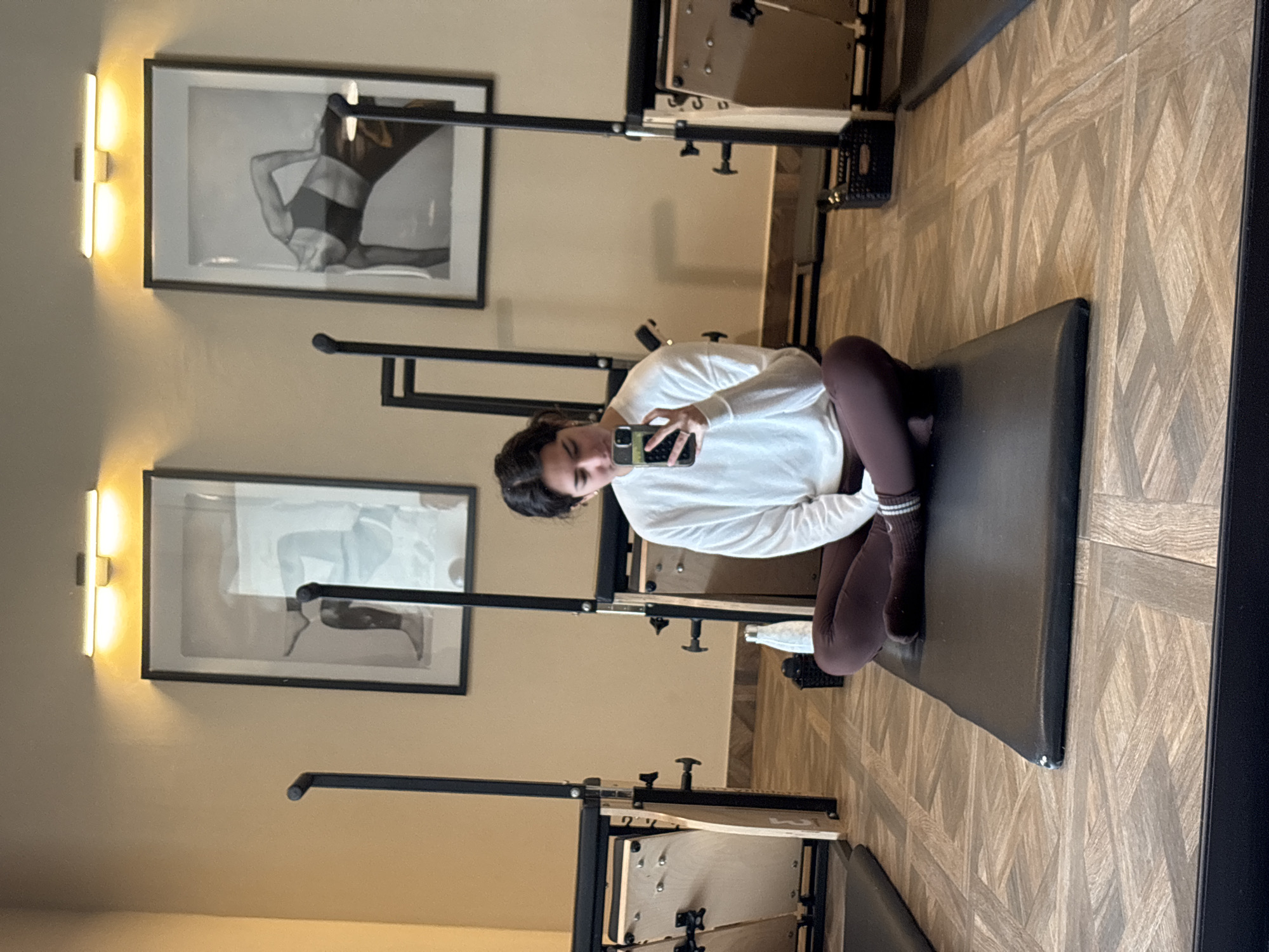 A woman is sitting on a pilates reformer, taking a selfie with her phone. She is wearing a white sweater and brown leggings. The reformer is in a studio with wood floors and beige walls. There are two framed black and white photographs hanging on the wall behind her. The studio is likely located in San Bernardino Tlaxcalancingo, Mexico.