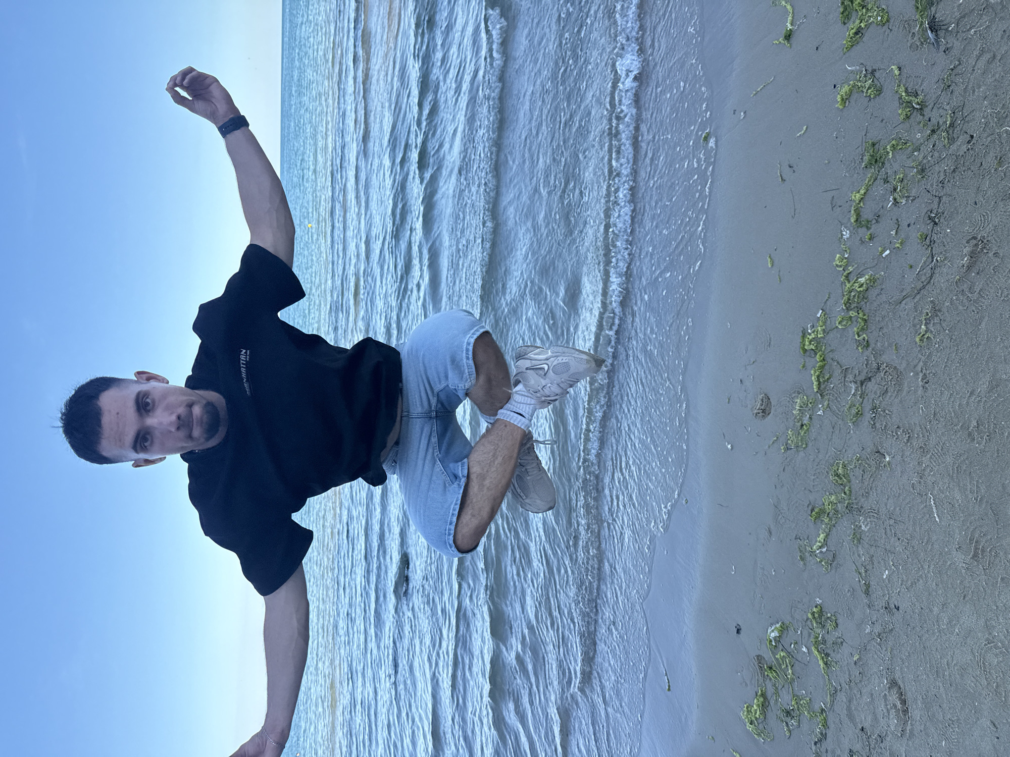 A young man with short dark hair and a goatee is captured mid-air, appearing to be jumping or dancing on a beach. He is wearing a black t-shirt with white text on the left chest, light blue denim shorts, white socks, and grey athletic shoes. His left arm is extended upwards, while his right arm is bent at the elbow, with his hand resting on his shorts. His legs are bent, and his body is positioned as if he is in motion.
The setting is a beach with the ocean in the background and the sandy shore in the foreground. The water is a calm blue, with gentle waves reaching the shore. The sand is light grey and has patches of green seaweed scattered across it. The sky is clear and bright blue, suggesting it is daytime, likely late afternoon or early evening given the soft light. The overall mood seems energetic and playful, reflecting the subject's dynamic pose.
The visible text on the man's t-shirt appears to be "MANHATTAN". The image is oriented vertically, with the subject positioned on the left side and the sea and beach filling the rest of the frame. The location context provided is Varna, Bulgaria.