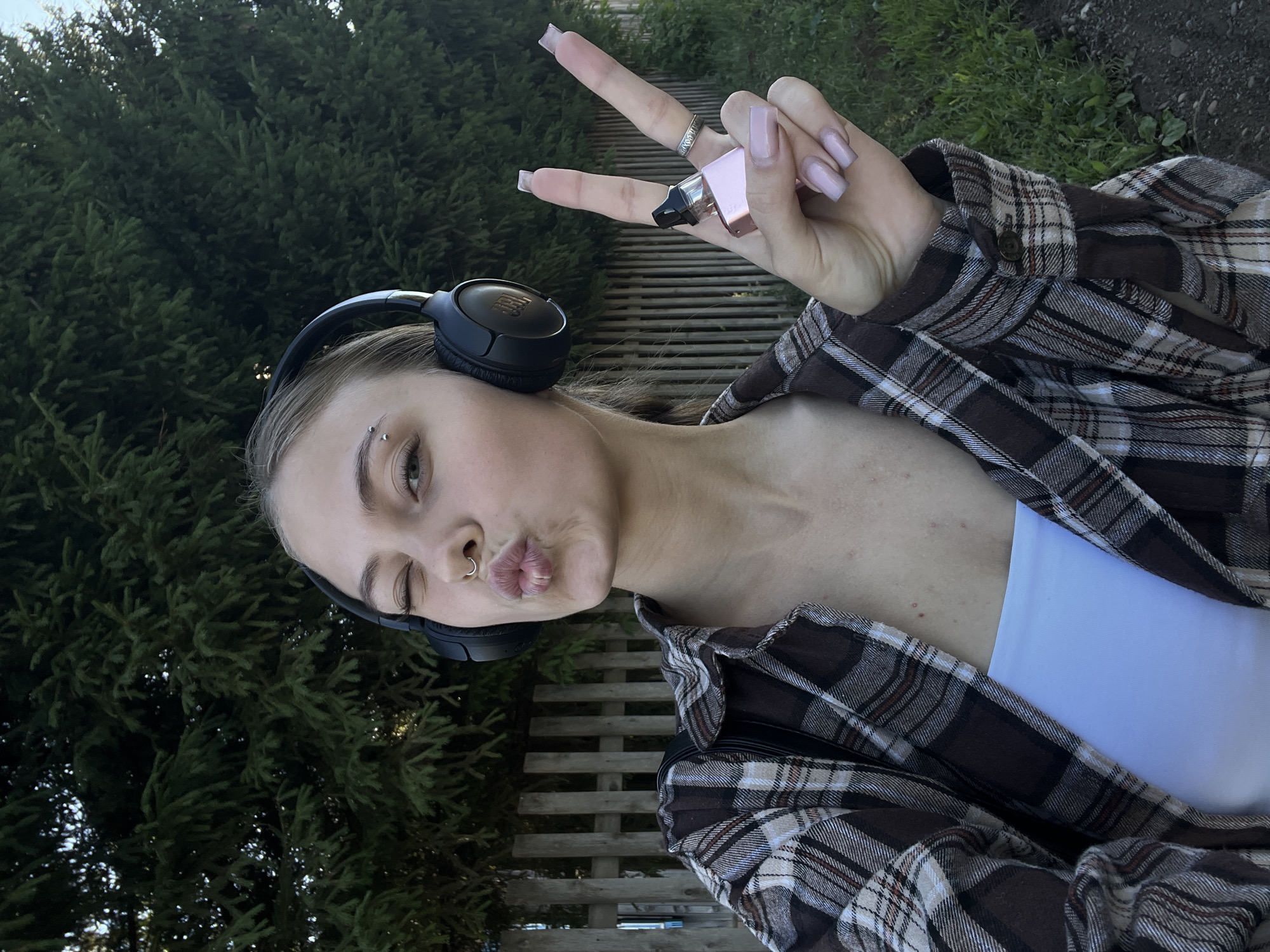 The image depicts a young woman with blonde hair, wearing black headphones, a brown and white plaid shirt open over a white tube top, and a nose ring. She is winking and puckering her lips, with her right hand held up in a peace sign, displaying a pink vape device and a ring. The background features a wooden fence and green trees, suggesting an outdoor setting. The lighting indicates it is daytime. No specific location in the city of Фоки, Russia can be identified from the provided image.